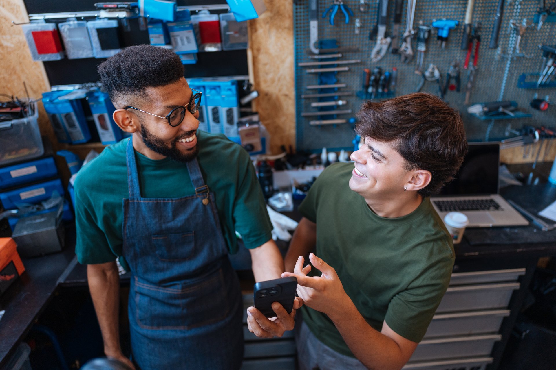 Two cheerful bicycle mechanics laughing and chatting while using a smartphone in a busy repair shop, showcasing teamwork and expertise