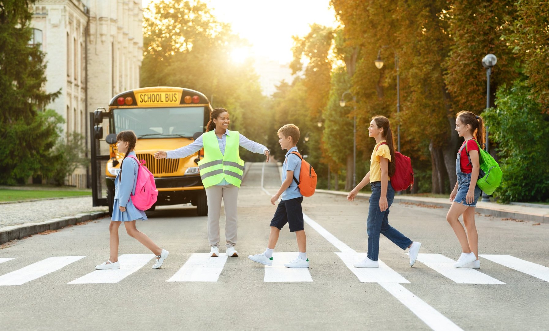 Group of children passing crosswalk on their way to school bus, diverse pupils cross the street with the crossing guard blocking traffic, boys and girls going home after classes