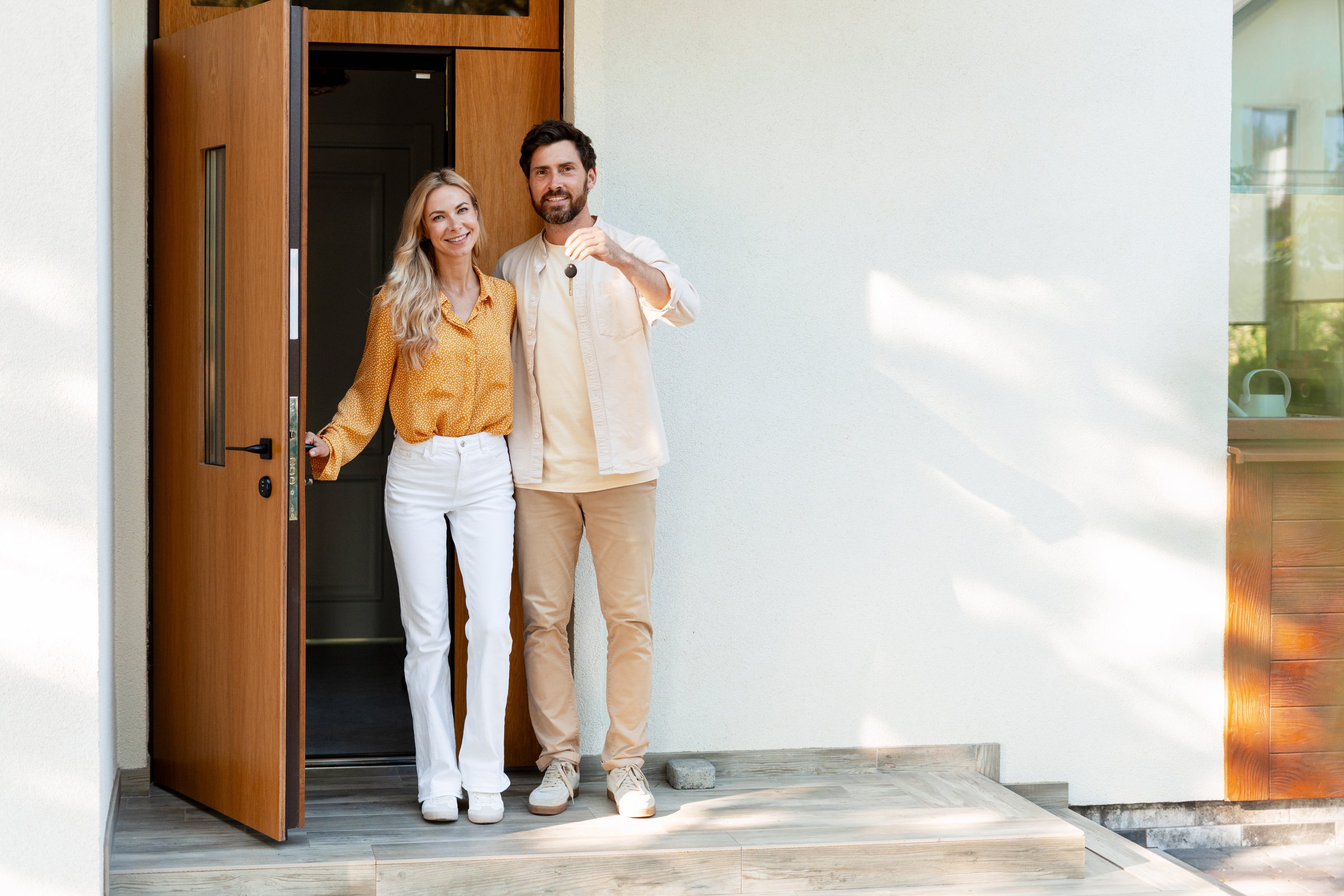 Happy couple is showing keys of their new home standing at the entrance door, smiling and looking at camera, real estate and accommodation concept