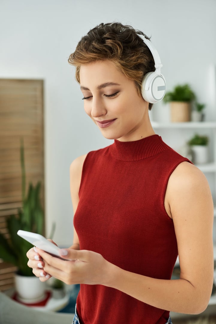 A young woman listens to music through headphones while checking her phone in a cozy home setting.