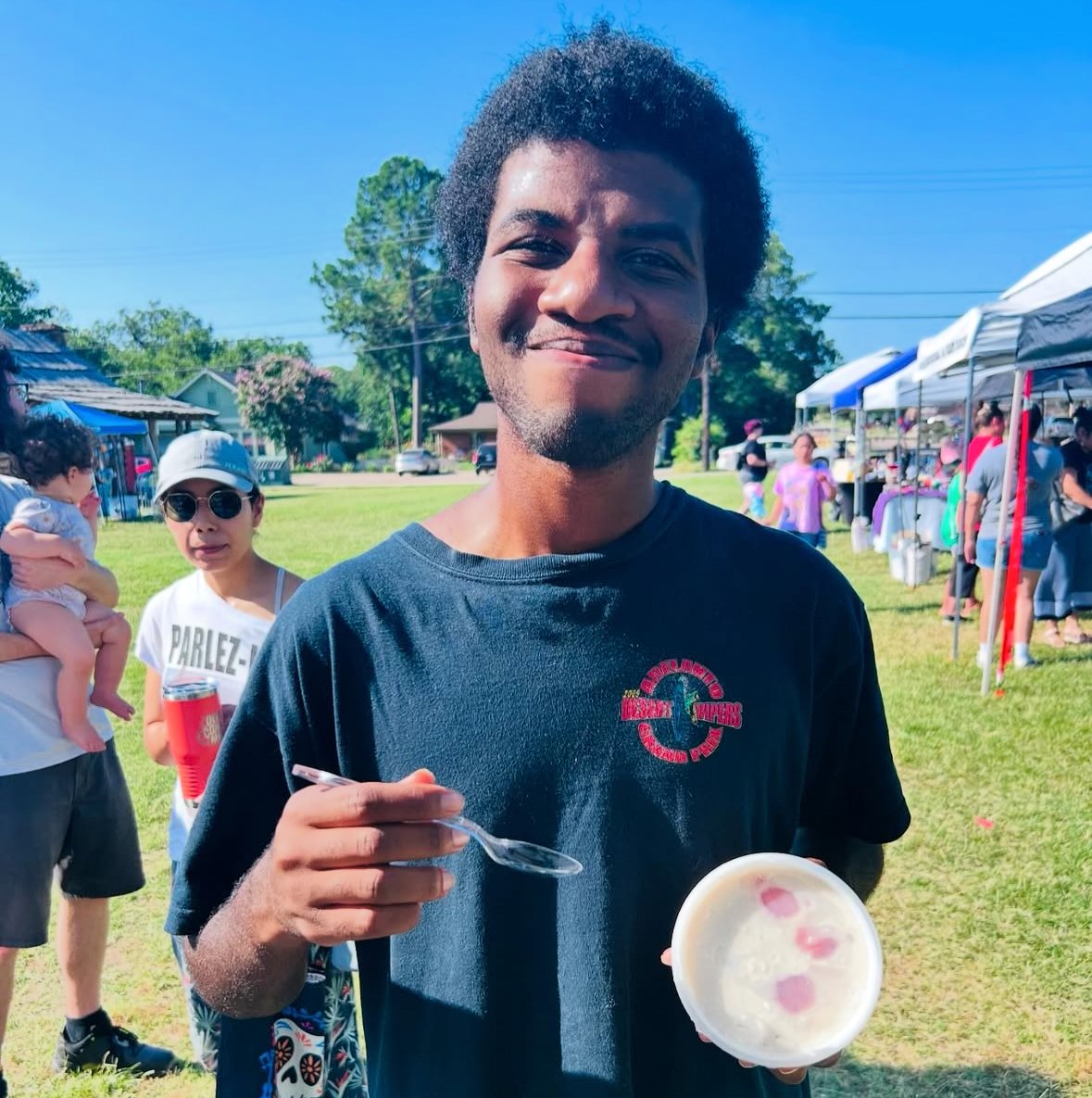 A smiling person holding a cup of shaved ice with cherries, standing outdoors at a market with tents and people in the background.
