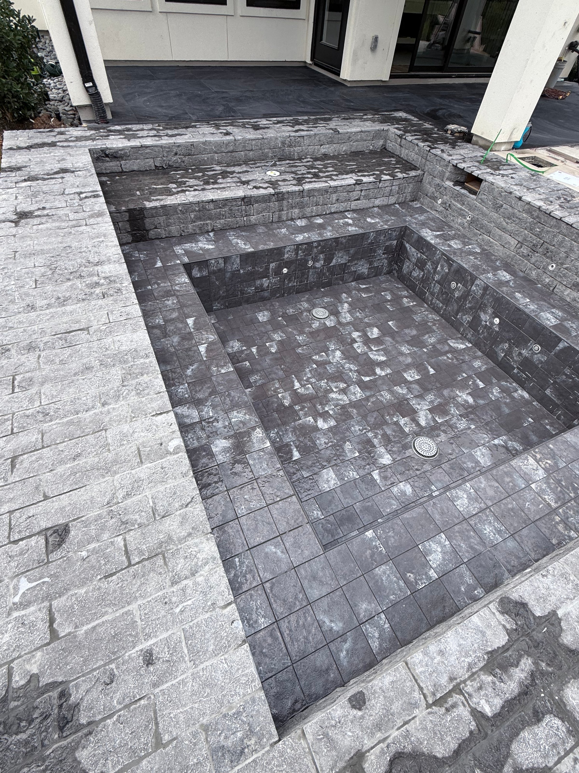 Empty rectangular hot tub with gray stone tiles in a backyard patio area.