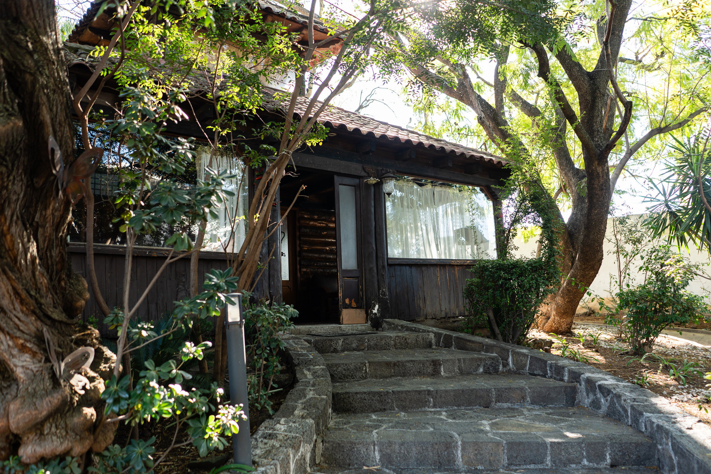 A rustic wooden cabin with a stone staircase, surrounded by lush trees and shrubs.