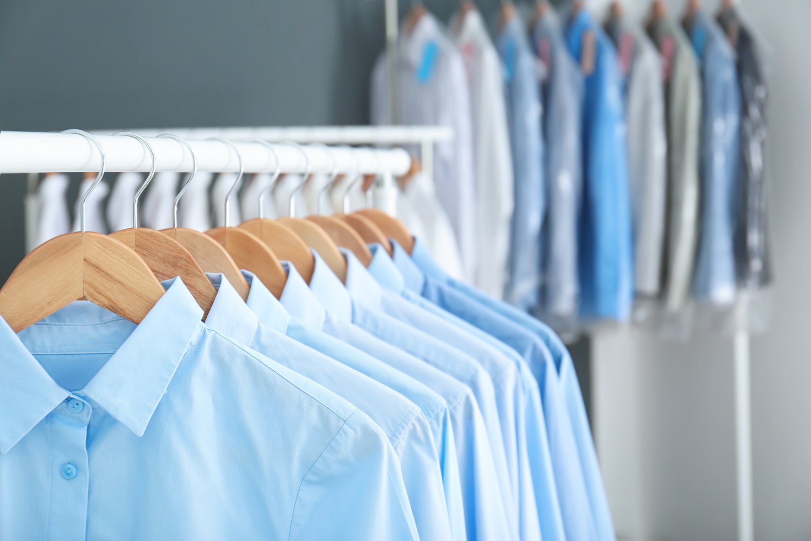 Close-up photograph of numerous light blue formal or business shirts hanging neatly on wooden hangers in a closet or dry cleaning rack, emphasizing professionalism and laundry care.