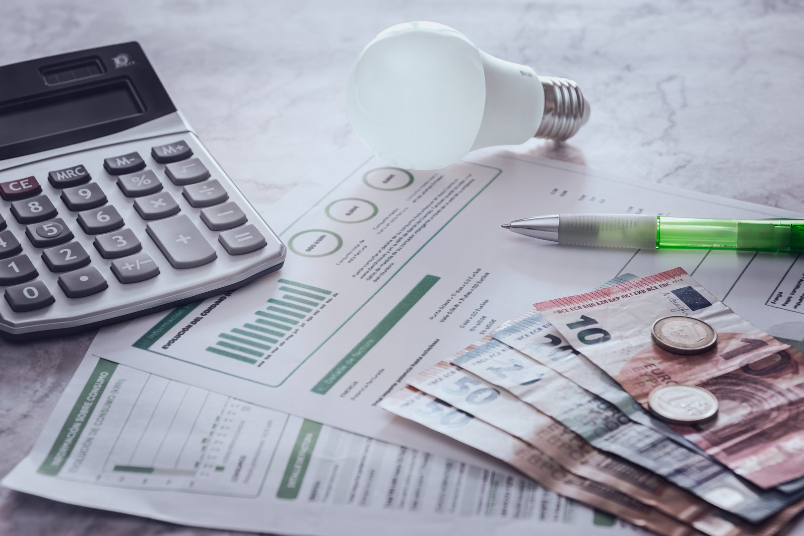 On a table several electric bills and on them an amount of money, a light bulb, a calculator and a pen.