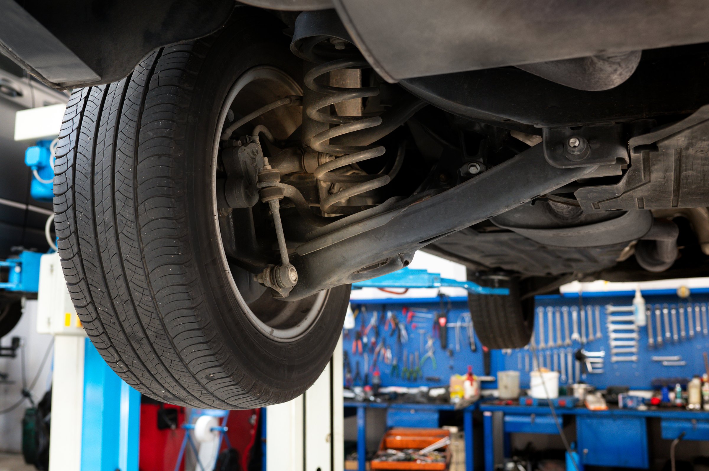 Bottom view of lifted car wheel with spring and shock absorber for repair in garage against neatly arranged various mechanic tools and instruments in daylight