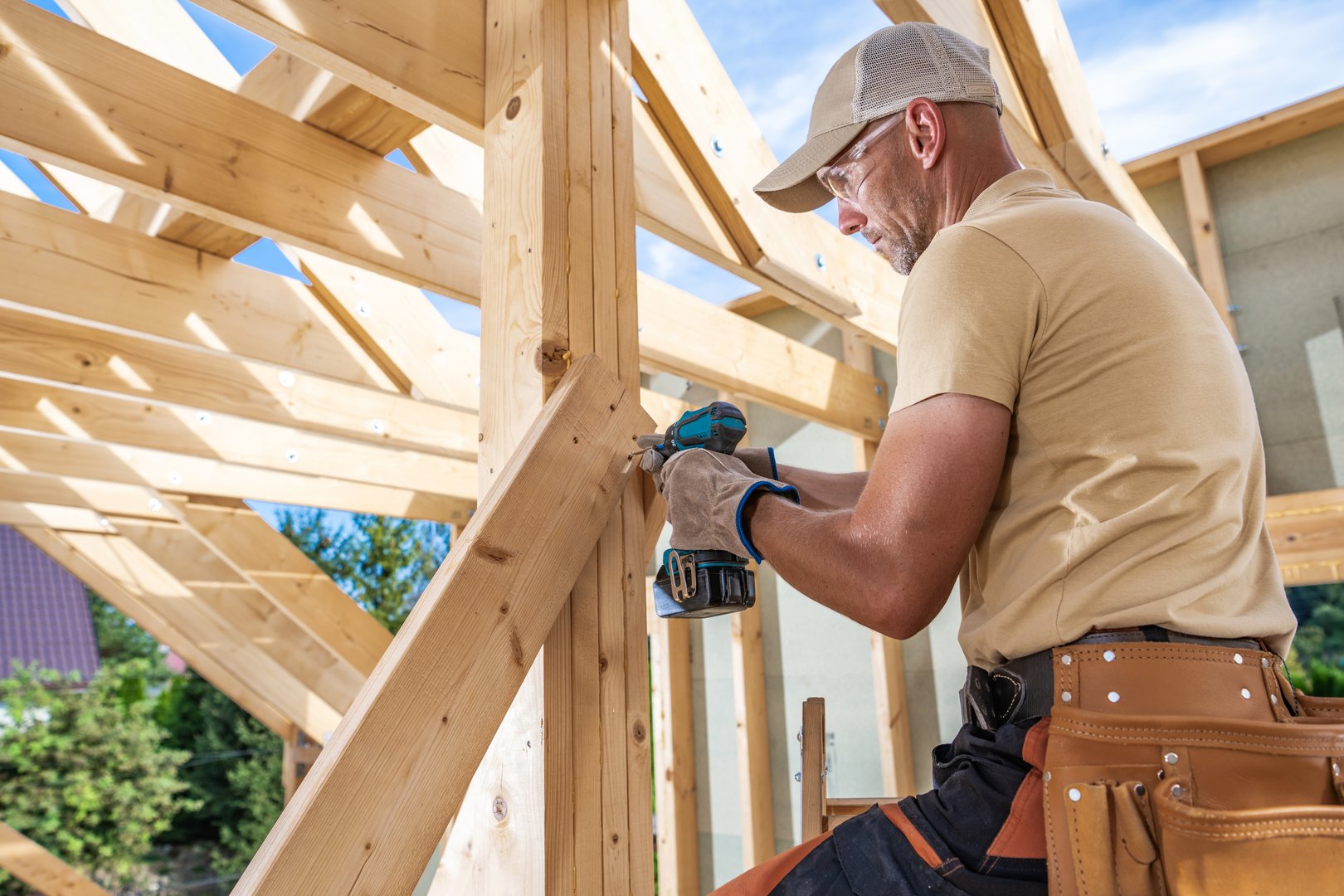 A carpenter uses a power drill to secure wooden beams while building a roof frame in a sunny residential area.