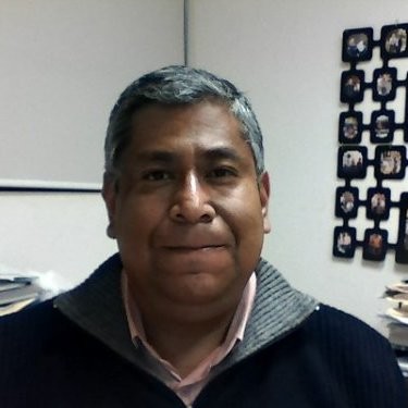 Man with short gray hair wearing a sweater, sitting in an office with a photo collage on the wall behind him.