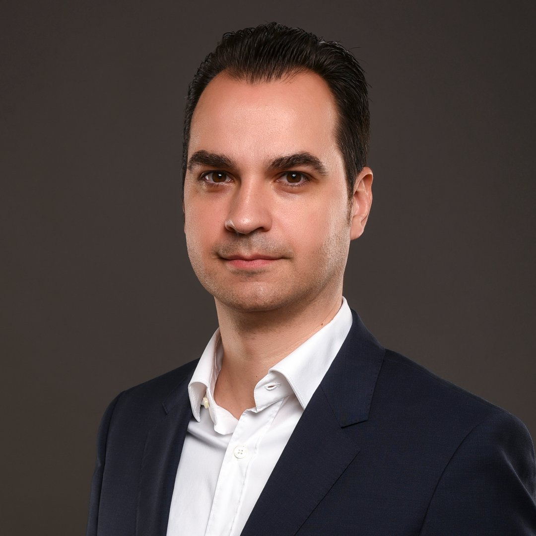 Portrait of a man in a dark suit and white shirt against a plain dark background.