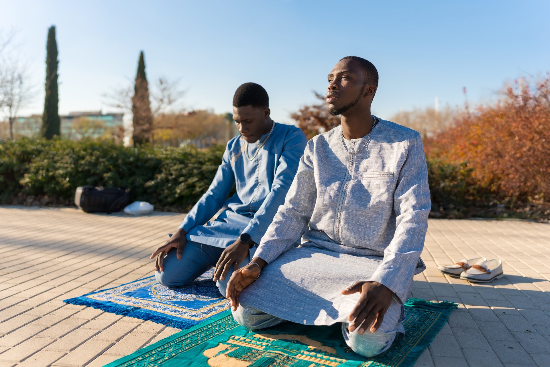 Two young senegalese men wearing traditional clothing are kneeling on prayer rugs, praying outdoors in observance of ramadan