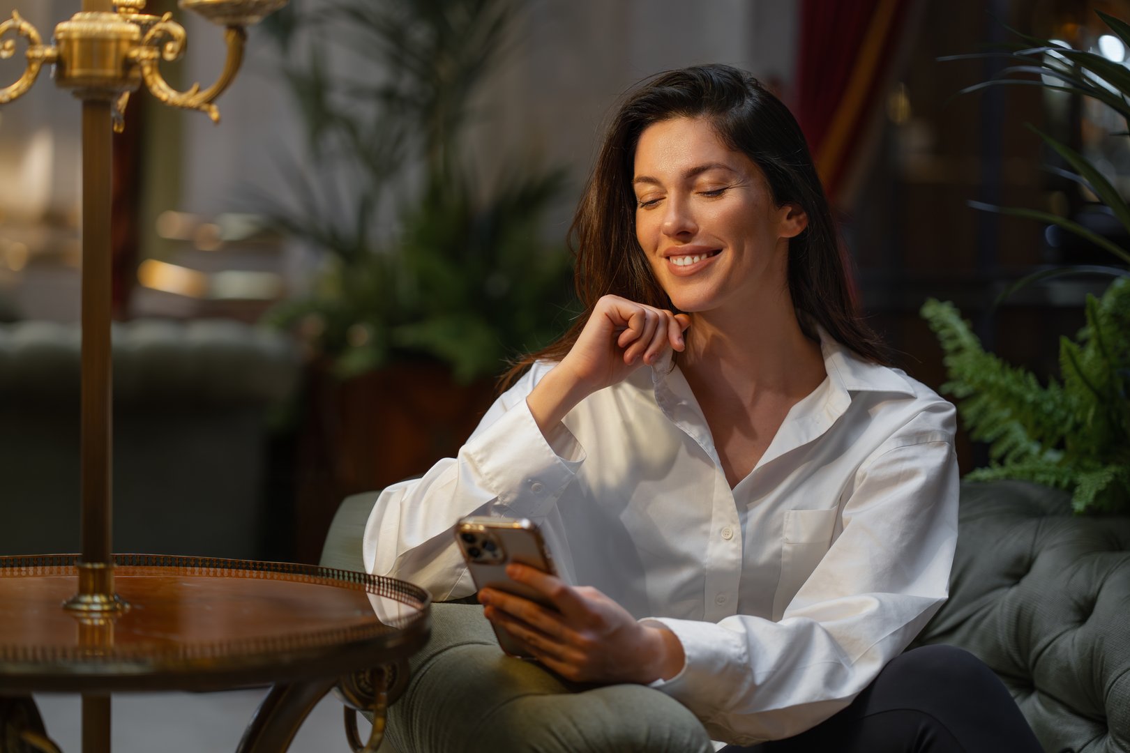 Smiling young businesswoman using smartphone sitting on cushioned chair in hotel lobby. Woman in white shirt and appears relaxed. Warm lighting create cozy atmosphere