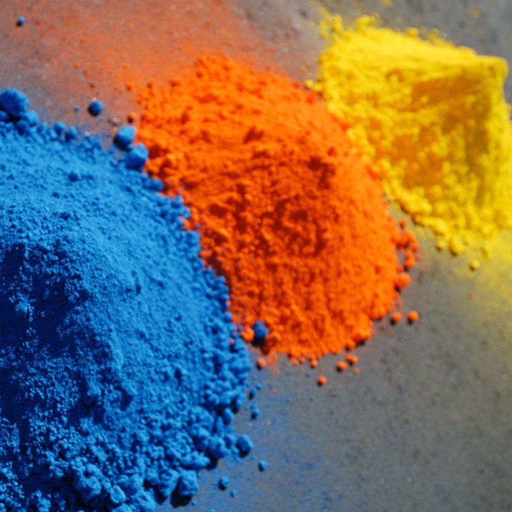 Piles of vibrant blue, orange, and yellow powders on a gray surface, side by side.