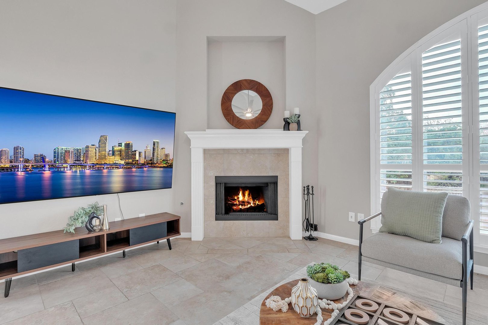 Modern living room with a fireplace, wall-mounted TV, decorative clock, and a large window with shutters.