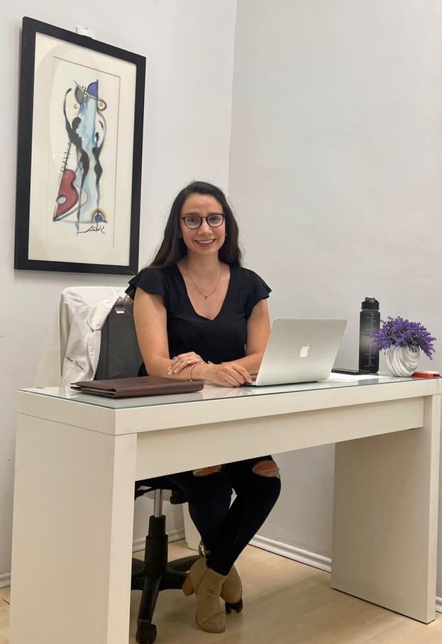 Woman sitting at a desk with a laptop, wearing glasses and a black top, with artwork on the wall and a vase of flowers.
