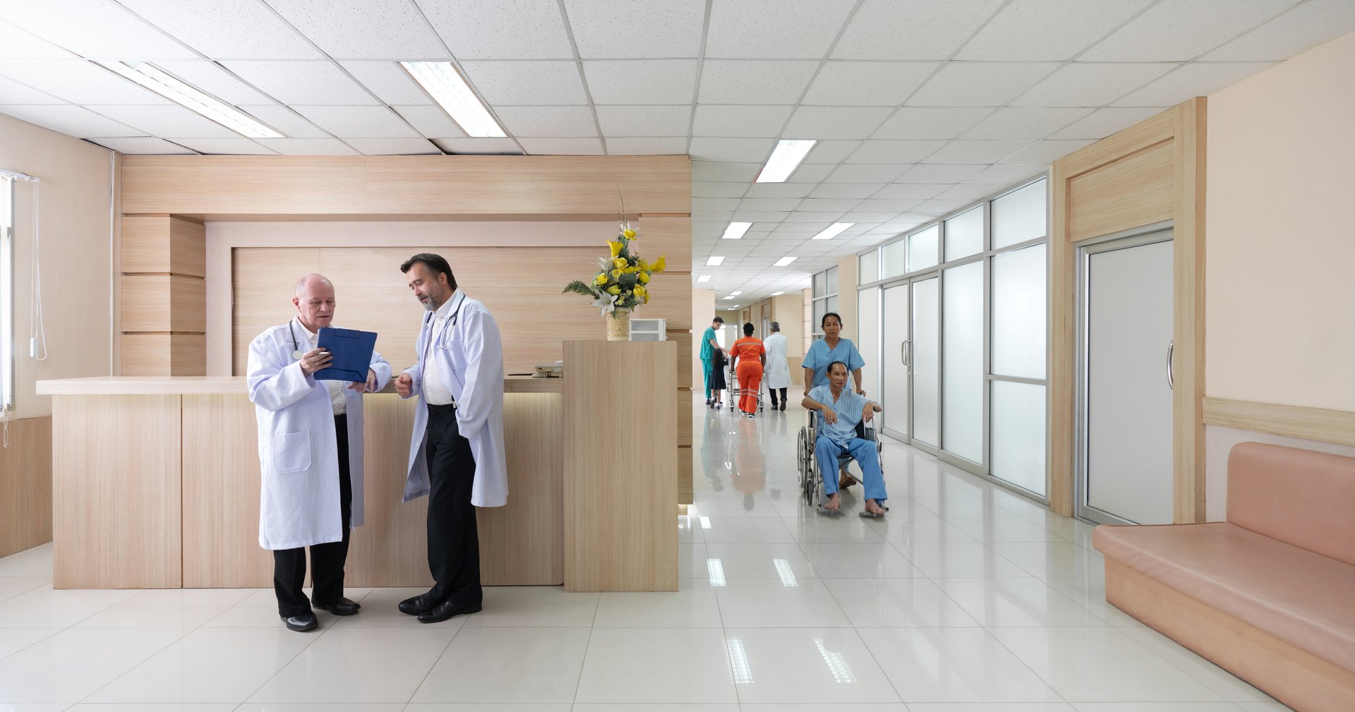 Two doctor in white coat review paperwork at a reception area while patient and nurse move through a bright, contemporary hospital corridor.
