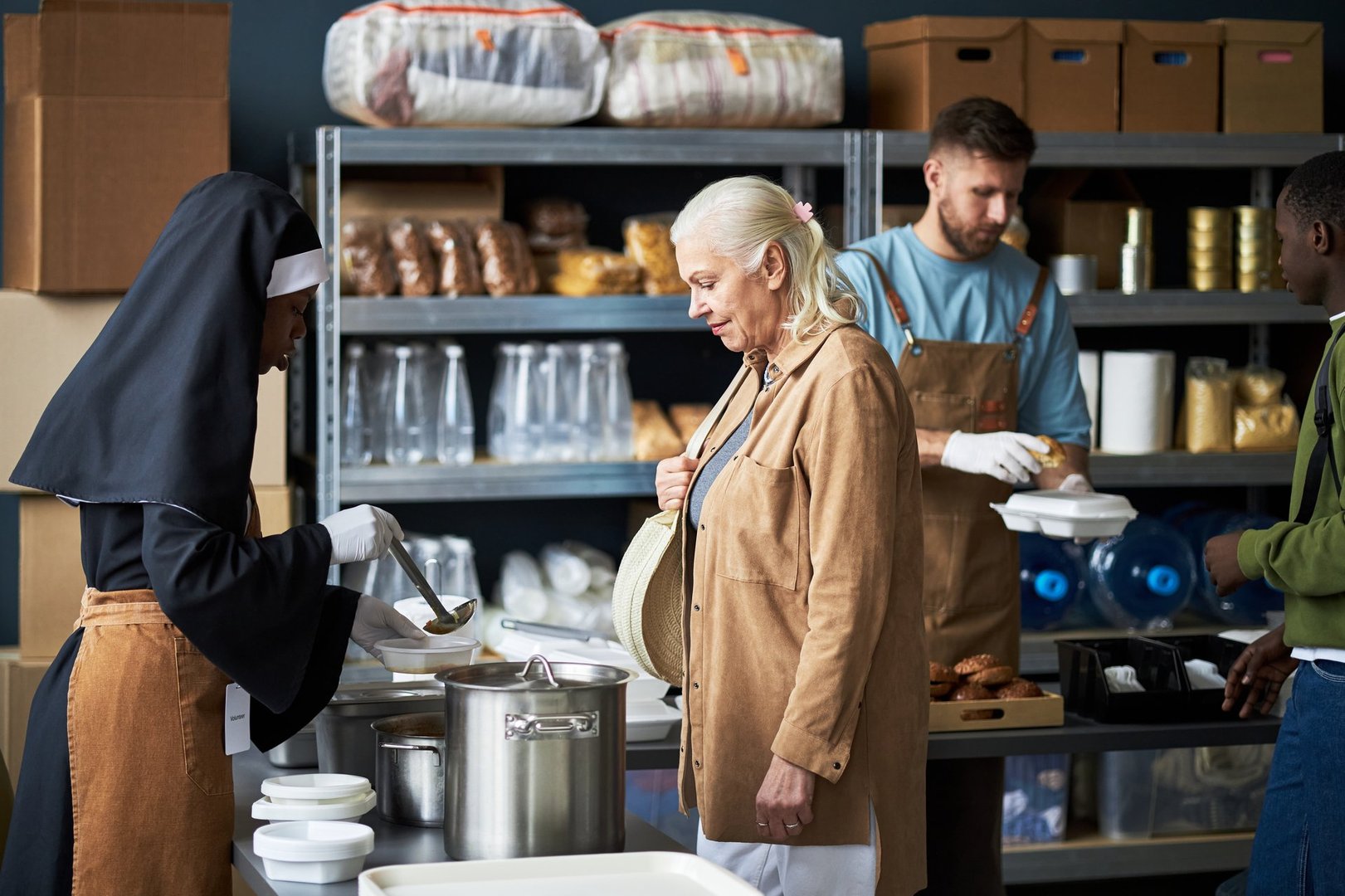 Volunteers of diverse backgrounds preparing meals in community kitchen setting Senior lady helping alongside nun and young men in background