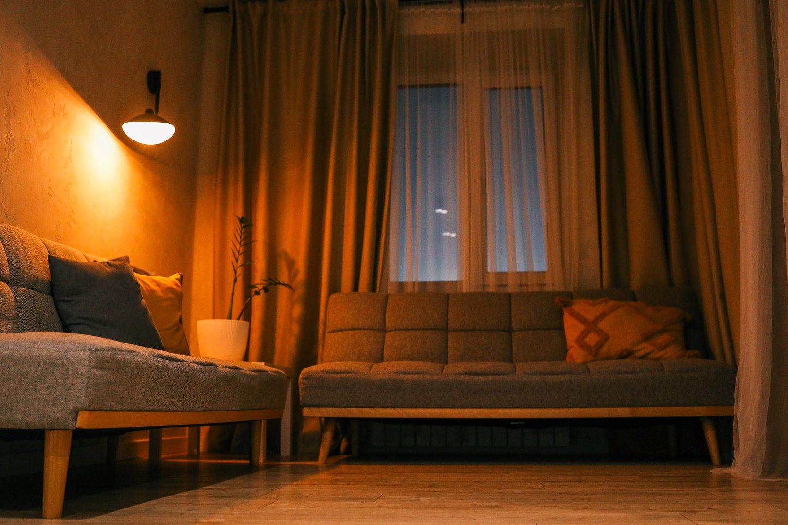 Cozy living room interior with warm lighting, grey sofas, decorative pillows, and a potted plant near the window during sunset.