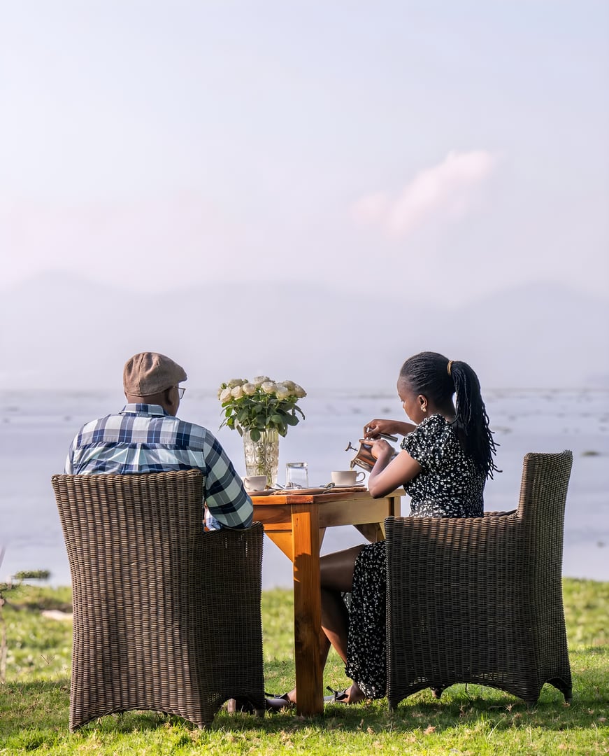 Two people sit at a lakeside table in wicker chairs, enjoying a meal with a view of water and mountains in the background.