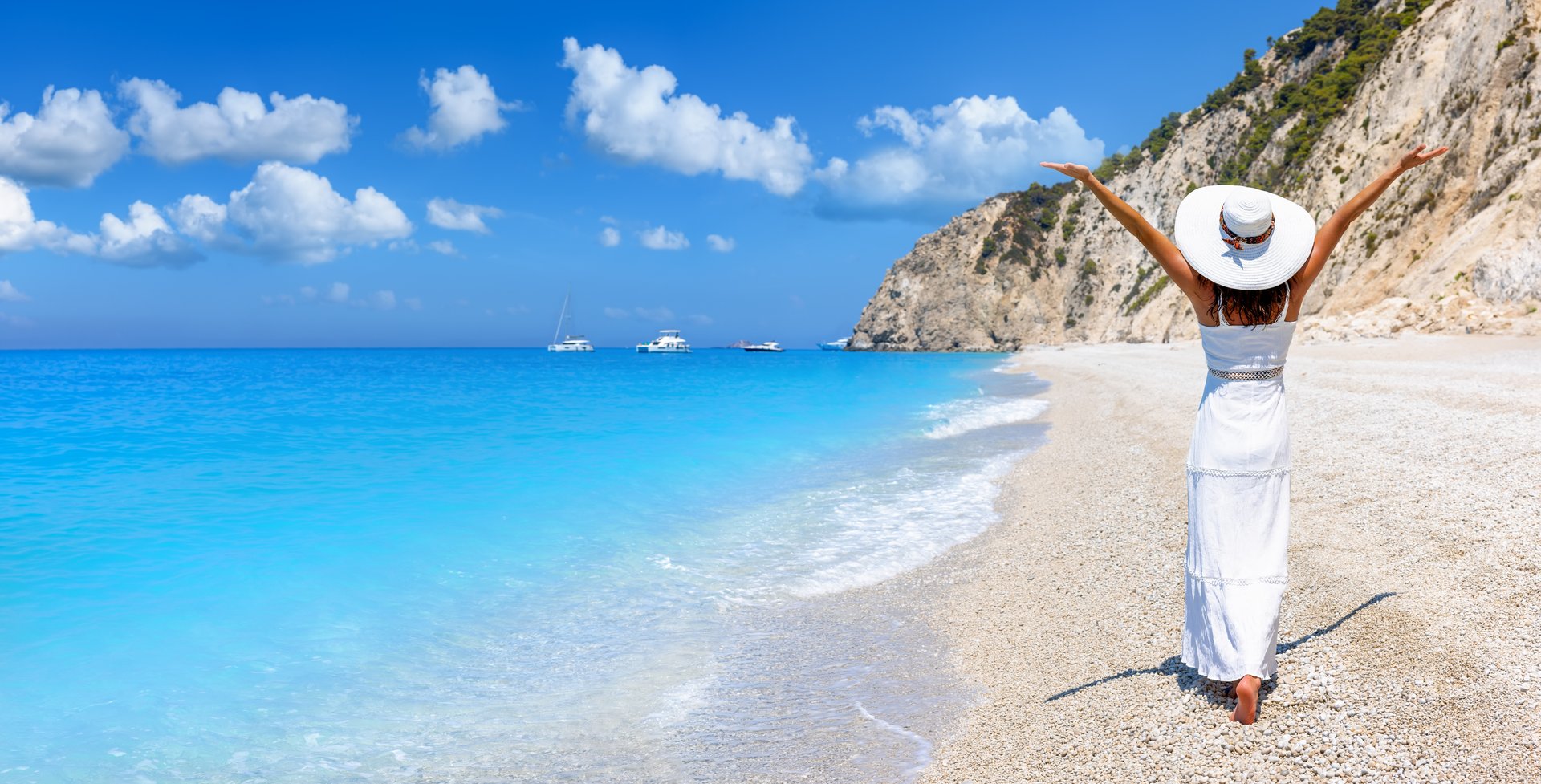 A happy tourist woman in a white summer dress walks down the famous Egremni Beach on the island of Lefkada, Greece