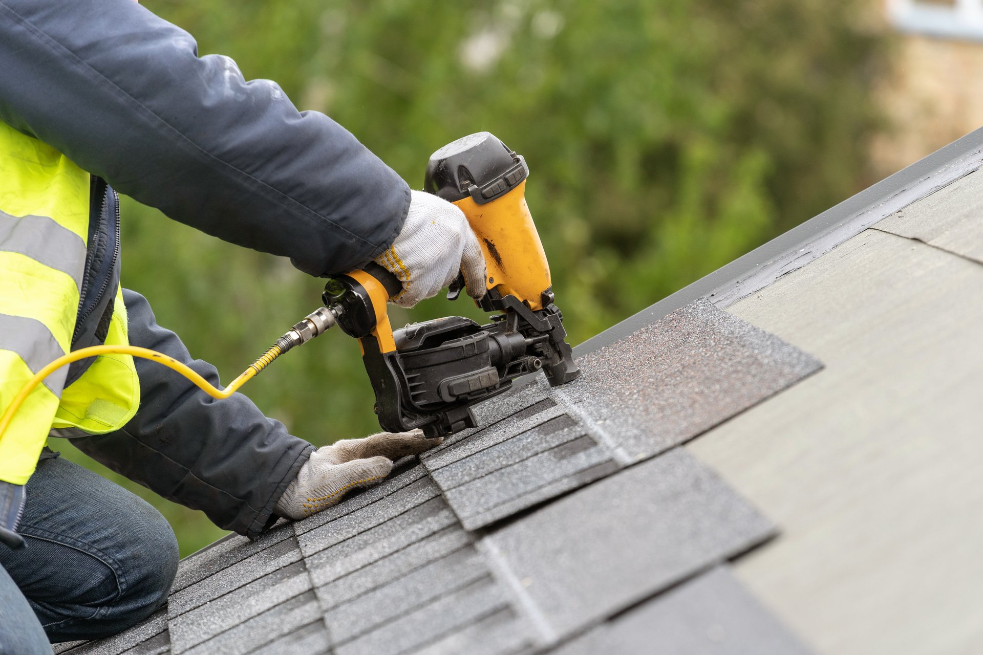 Roofer worker in uniform using nail gun installing shingles