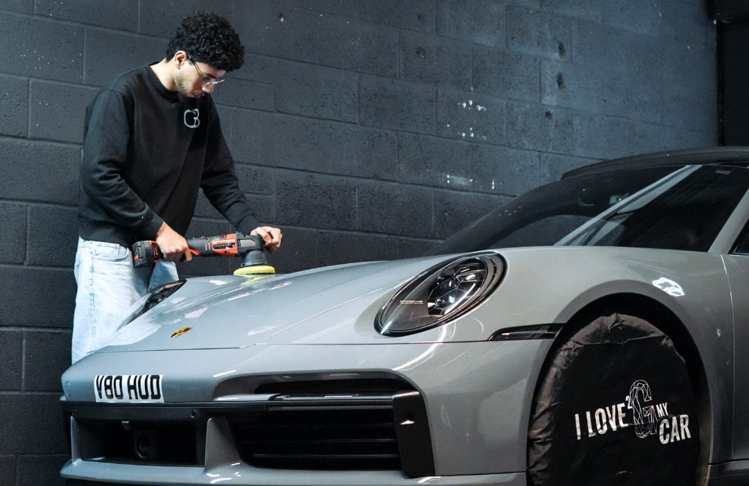 Person polishing a gray sports car in a garage, using a buffer on the hood. Car has a cover reading I LOVE MY CAR.