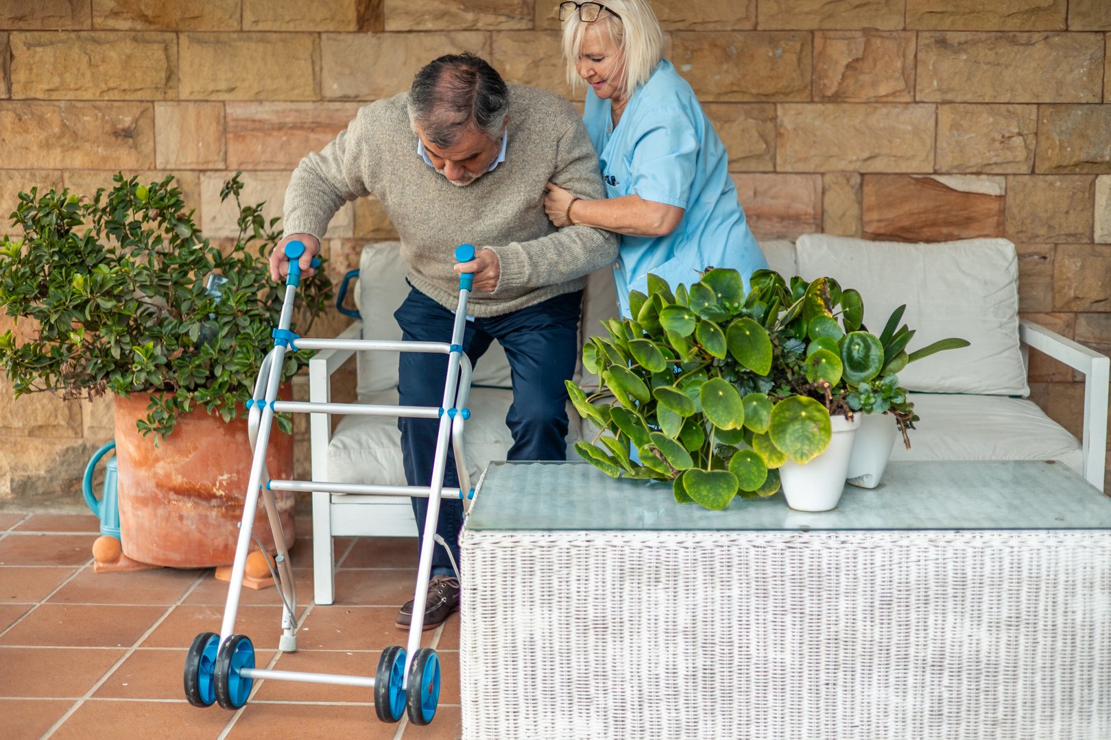 Nurse helps an elderly man to stand up using a walker in the garden of a rehabilitation center, providing support and encouragement during his recovery