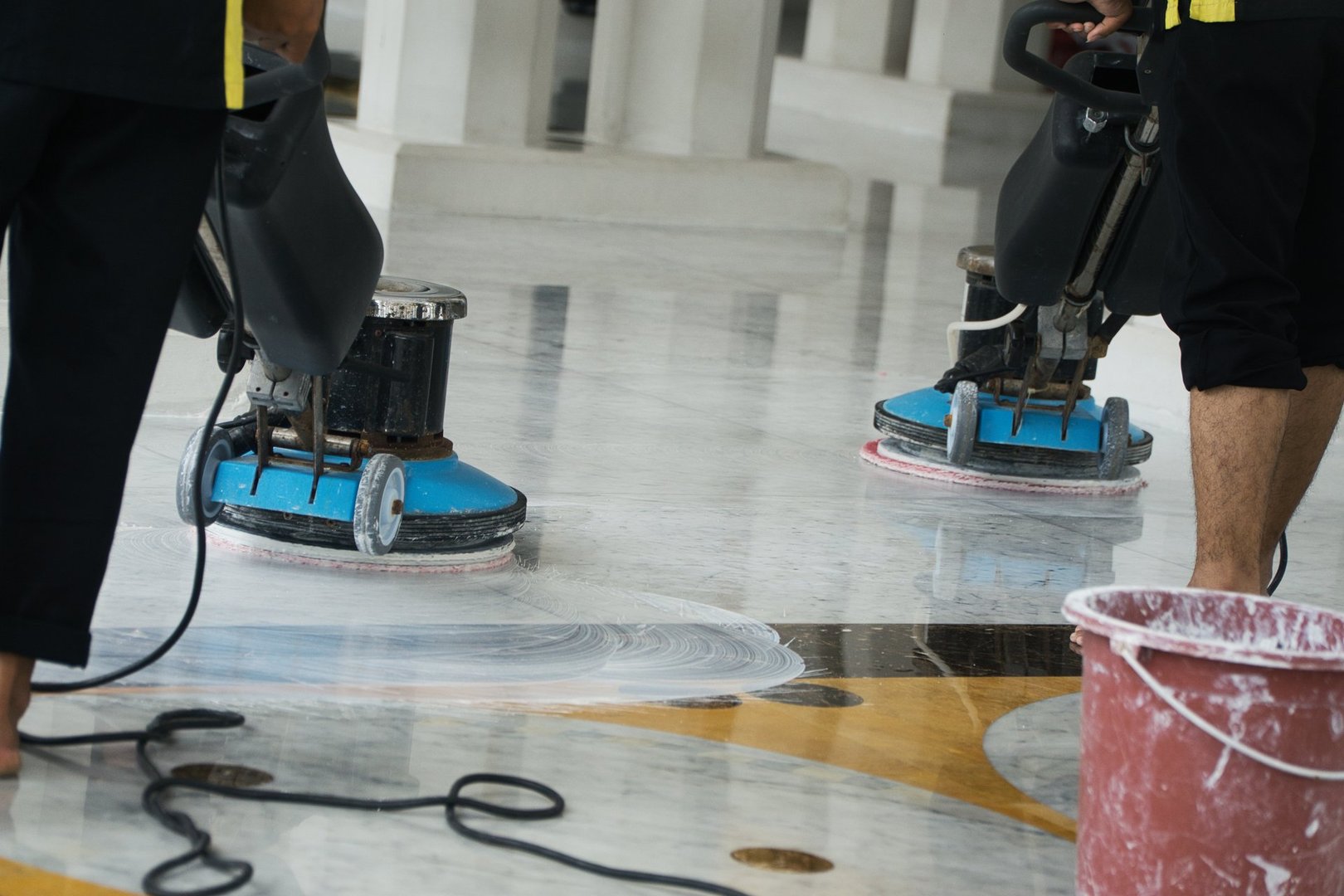 Two workers operate buffing machines to thoroughly clean a polished marble floor, ensuring it maintains its professional and spotless appearance. Equipment and accompanying cleaning supplies are visible, emphasizing a high standard of cleanliness