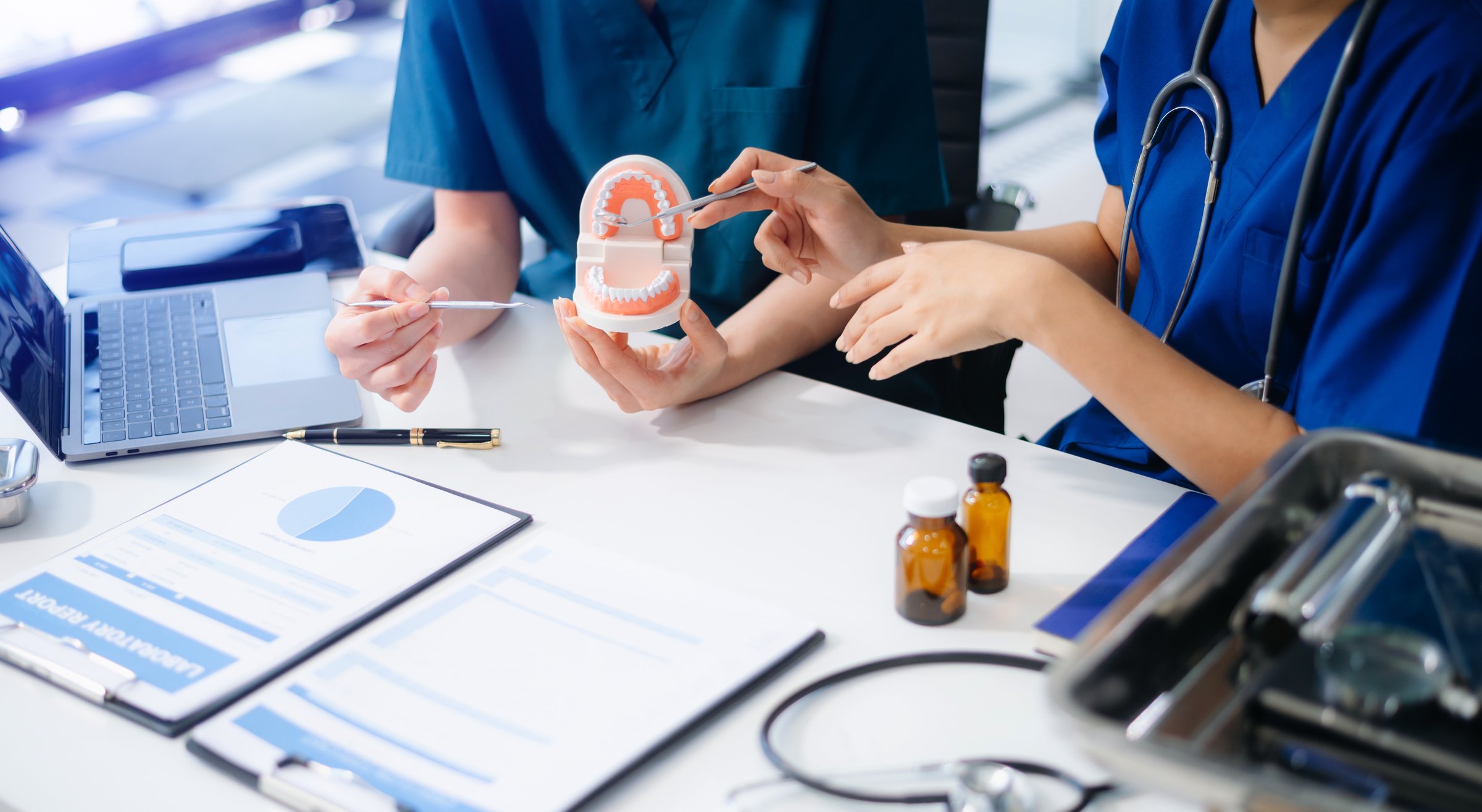 Two dentists in uniform type dental history on laptop with X-ray and tooth model on desk. Modern oral care, teamwork, and digital record in dental clinic.