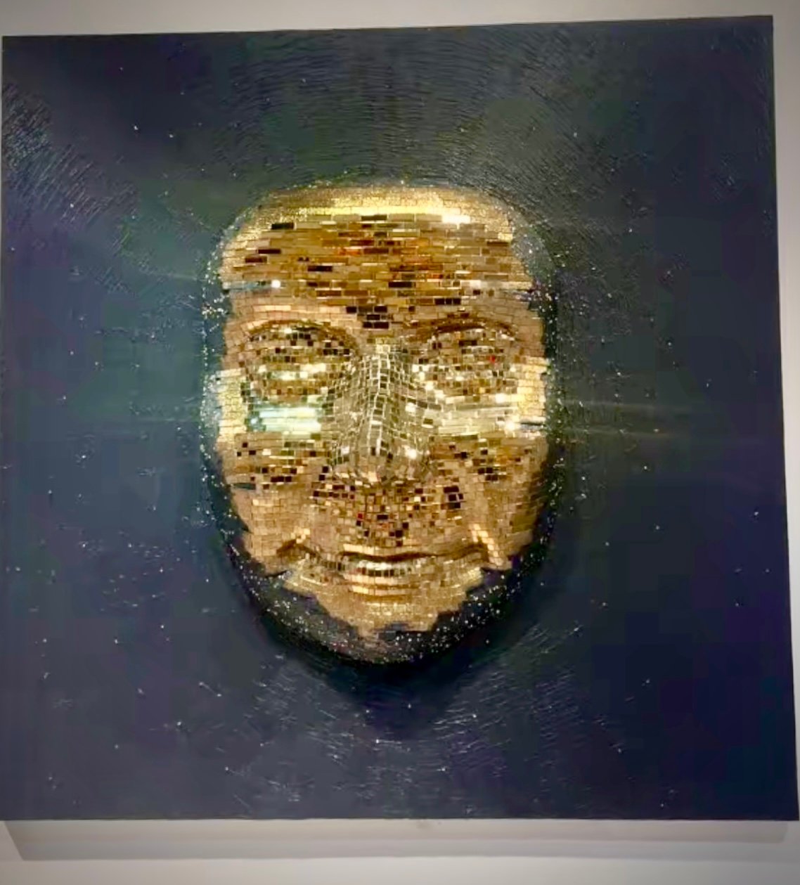 A mosaic art piece depicting a golden face with intricate details against a dark background, creating a shimmering effect.