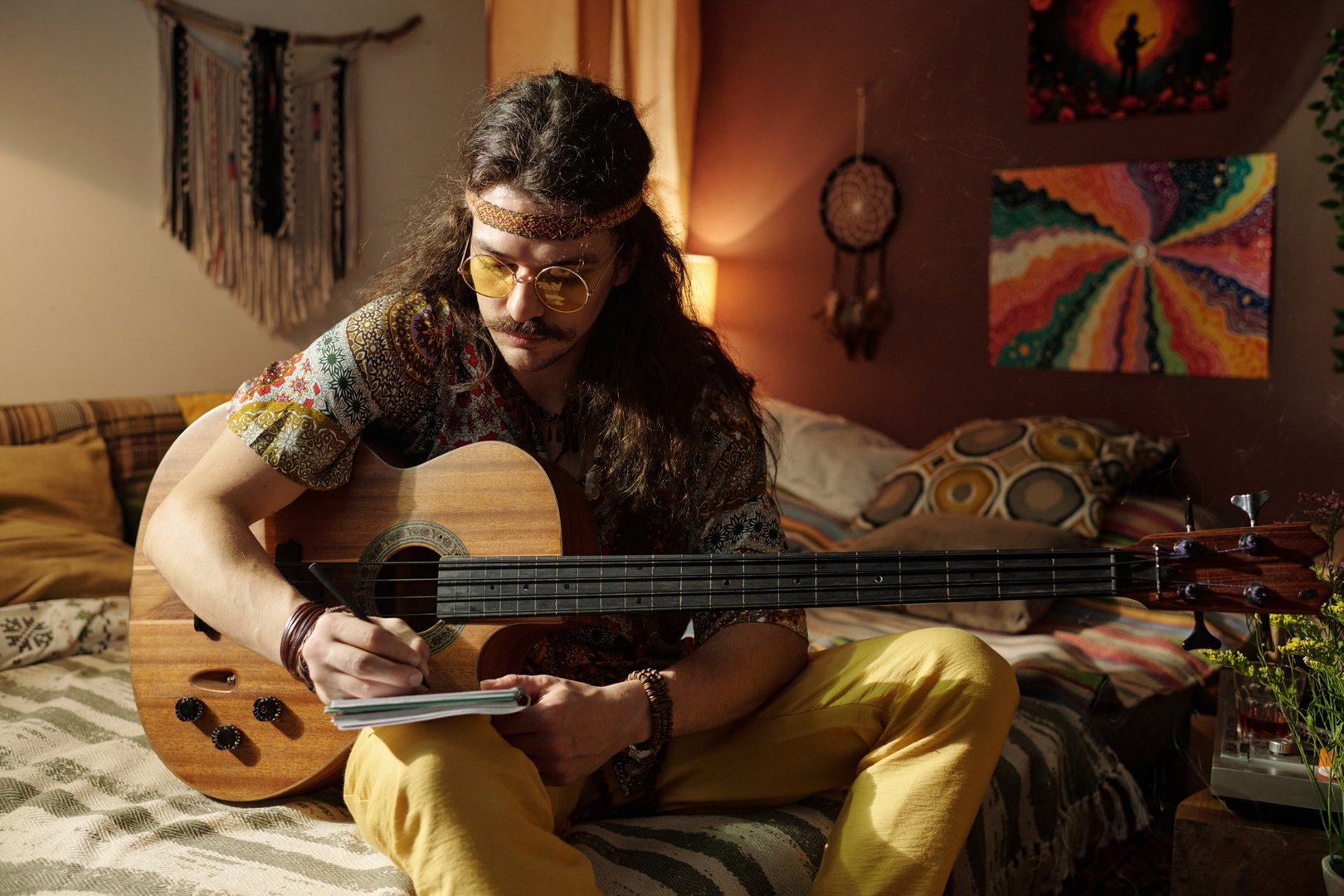 Musician sitting on bed with guitar, concentrating on writing music in cozy room with an artistic backdrop including a colorful painting and room decor