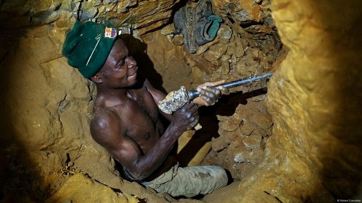 A miner in a narrow underground tunnel uses a tool to extract minerals. He wears a green beanie and is shirtless.