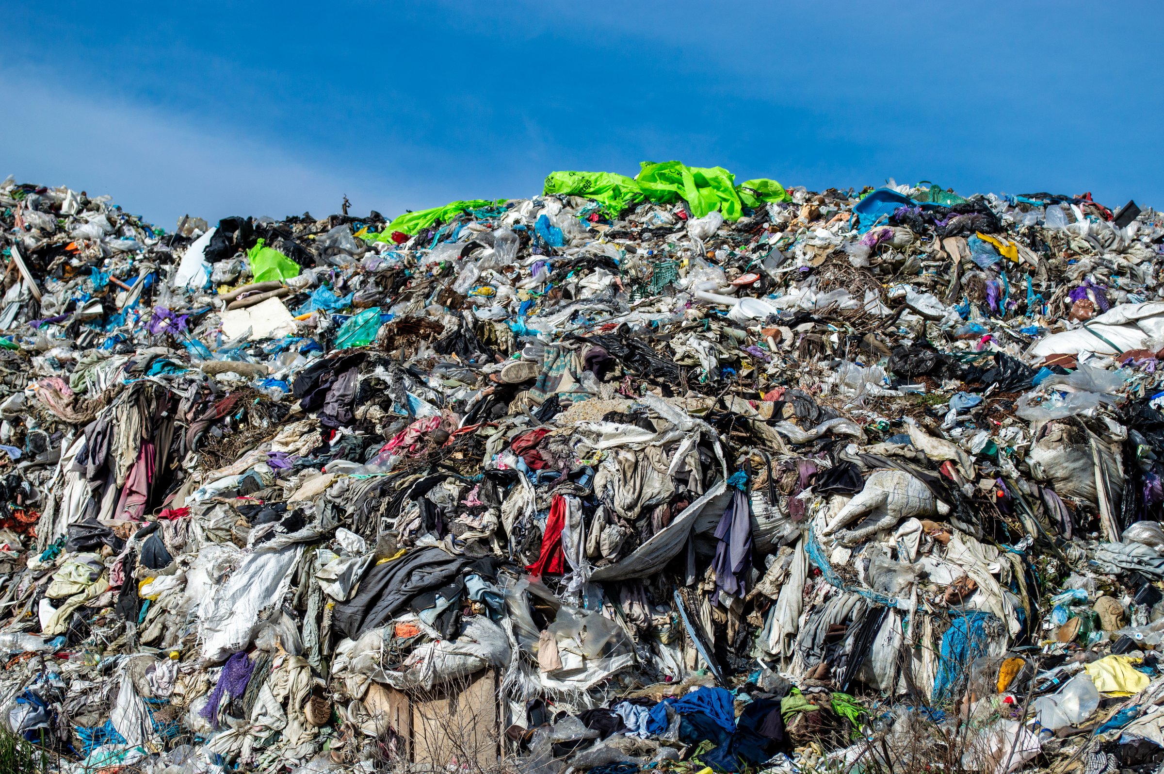 A vast accumulation of discarded clothing and fabrics fills the landfill, highlighting the issue of textile waste under bright sunlight.