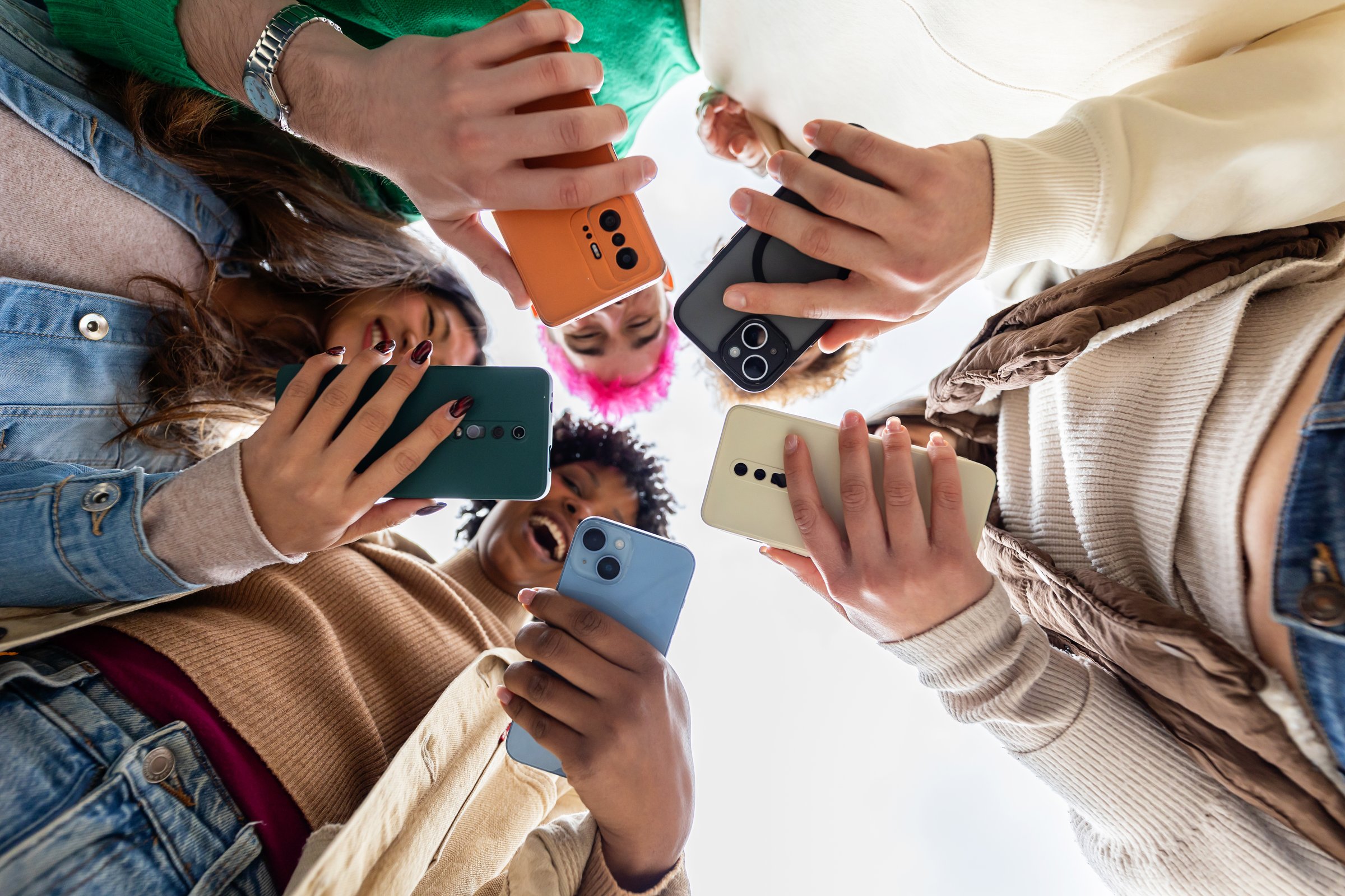 Young group of happy young student teenage friends having fun using mobile phone together outdoor, watching social media content or playing video game on smart phone device. Technology lifestyle