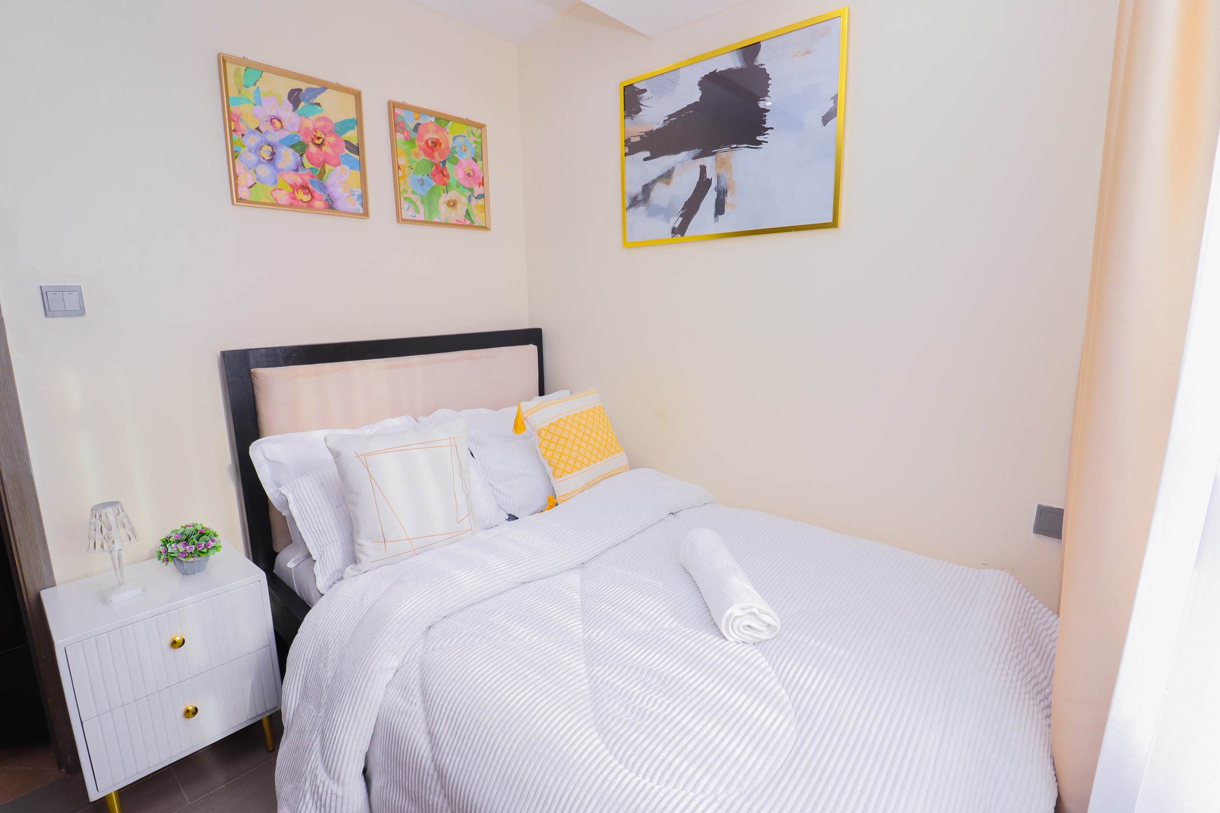 Cozy bedroom with a neatly made bed, white bedding, decorative pillows, a nightstand, and colorful wall art.