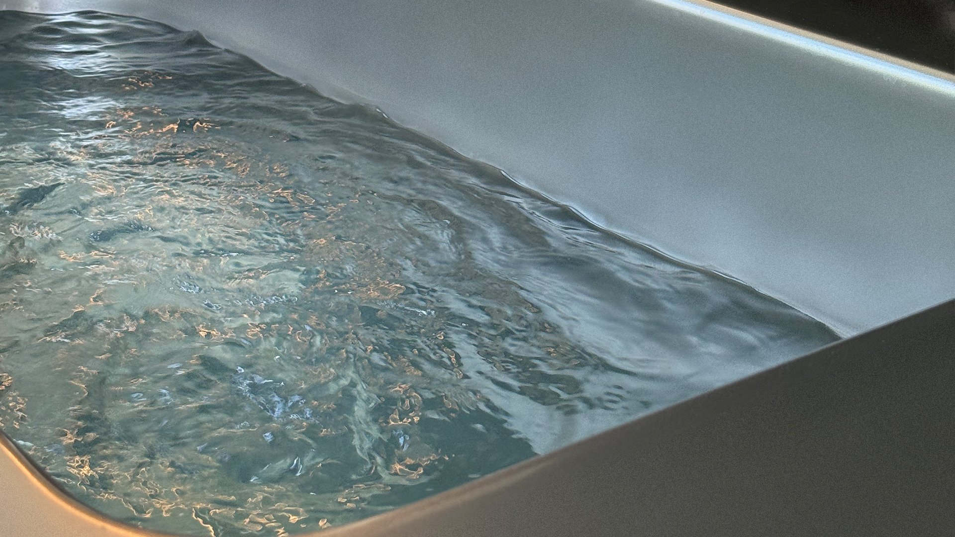 A partially filled bathtub with clear, gently rippling water, reflecting soft lighting from above.