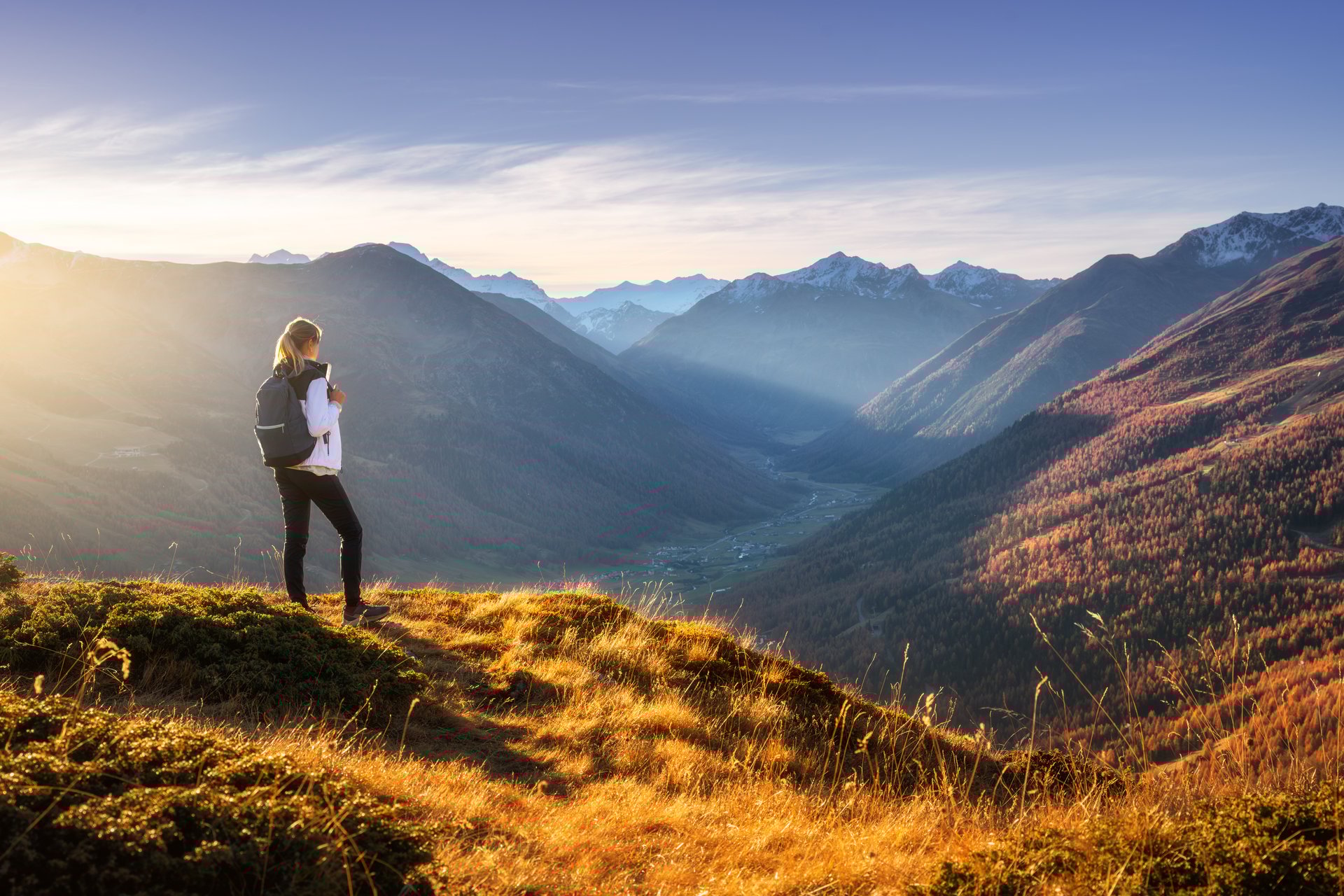 Young woman with backpack standing on grassy mountain ridge with view of valley with high mountain peaks during golden hour sunset in autumn. Hiking in Italy, Livigno. Girl in Alps in fall. Trekking