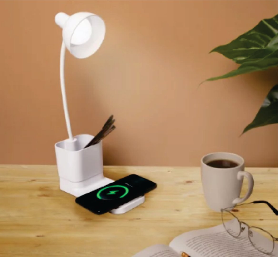 Desk with a white lamp, charging phone, cup of coffee, and plant on a wooden surface. Glasses and pens are also visible.