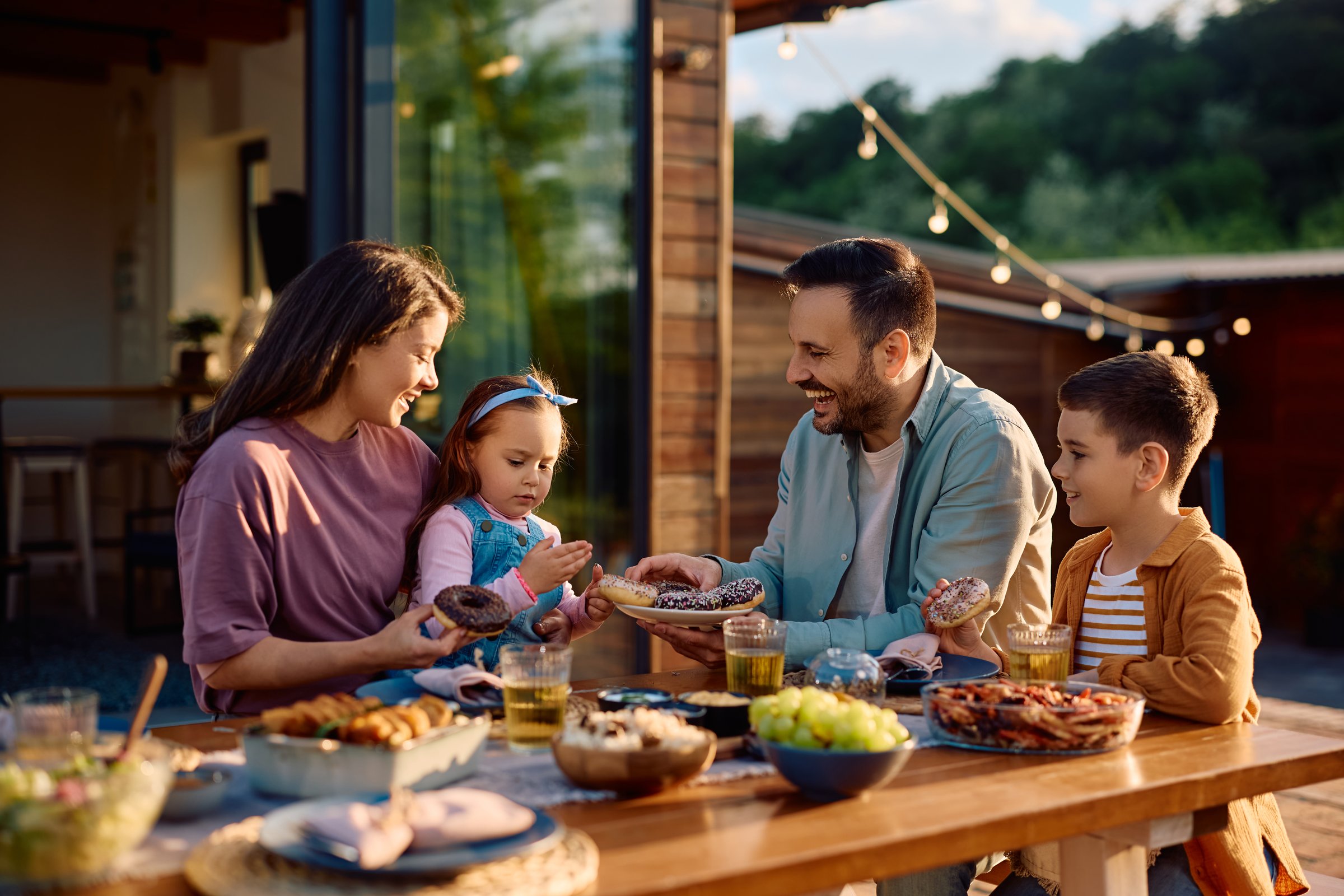 Happy parents and their kids eating donuts during family picnic day in their backyard.