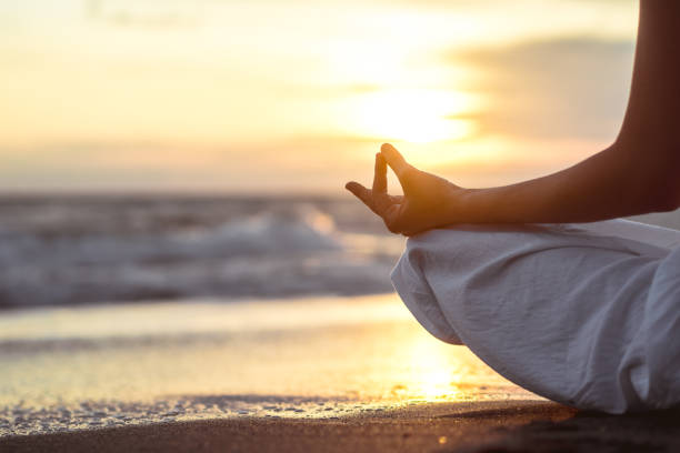 Person meditating on a beach at sunset, sitting cross-legged with hand in a mudra position, waves in the background.