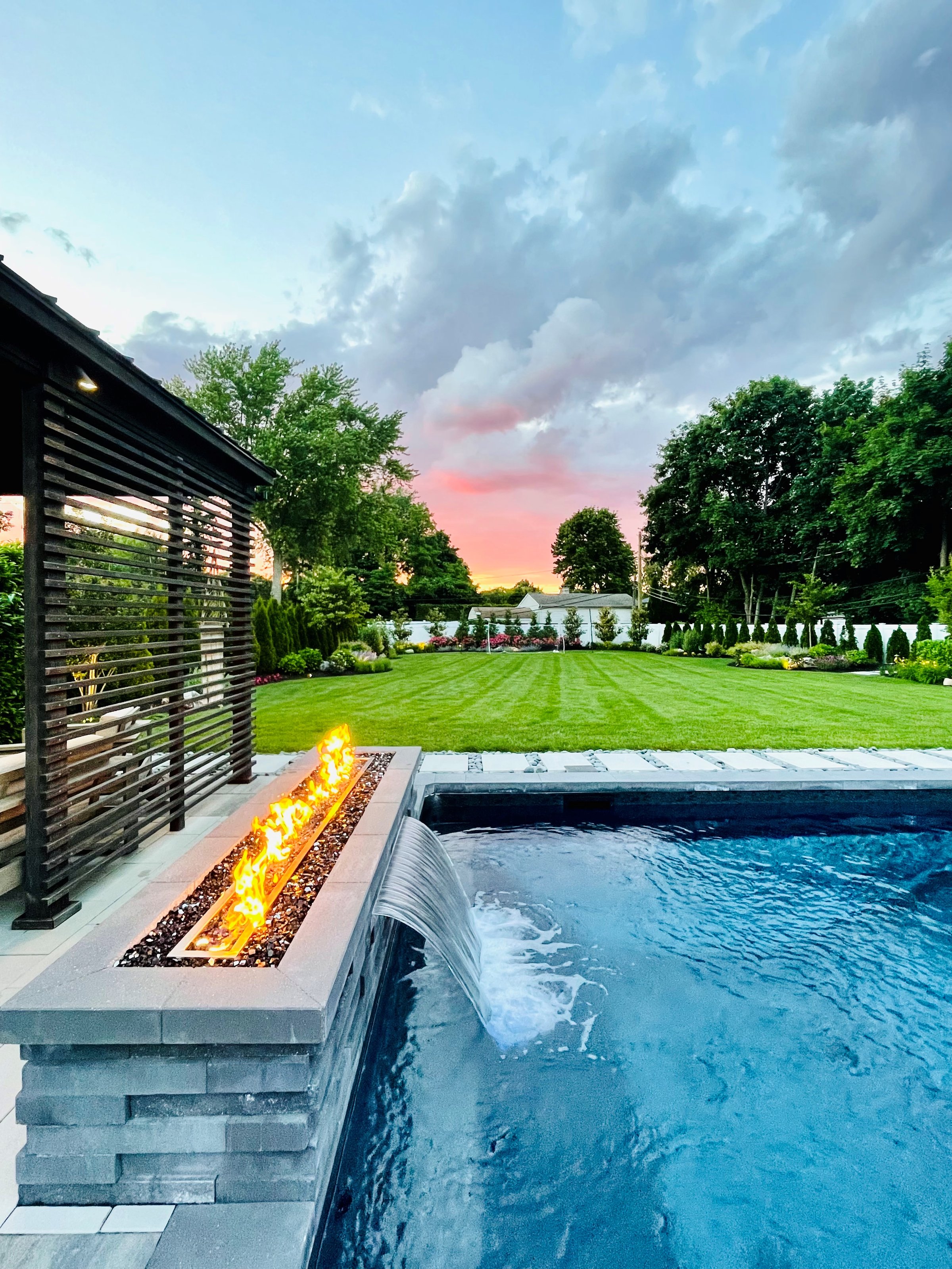 High-end, NY outdoor living space, a backyard patio with pergola, pavilion, cabana, outdoor rug,  tequila bar, dining set, couch, television, pool with waterfall, umbrella, fire pit and stunning grey patio. Modern vibe. Surrounded by lush cottage style gardens, flowers, shrubs and trees.  Stunning entertainment space to be used all year round and to enjoy in the summer time.  amazing night display of accent lighting lights up the property in color - here is shows vivid flames from firepit.  Cool off in the pool, stay warm by the fire.  Fire and water display.