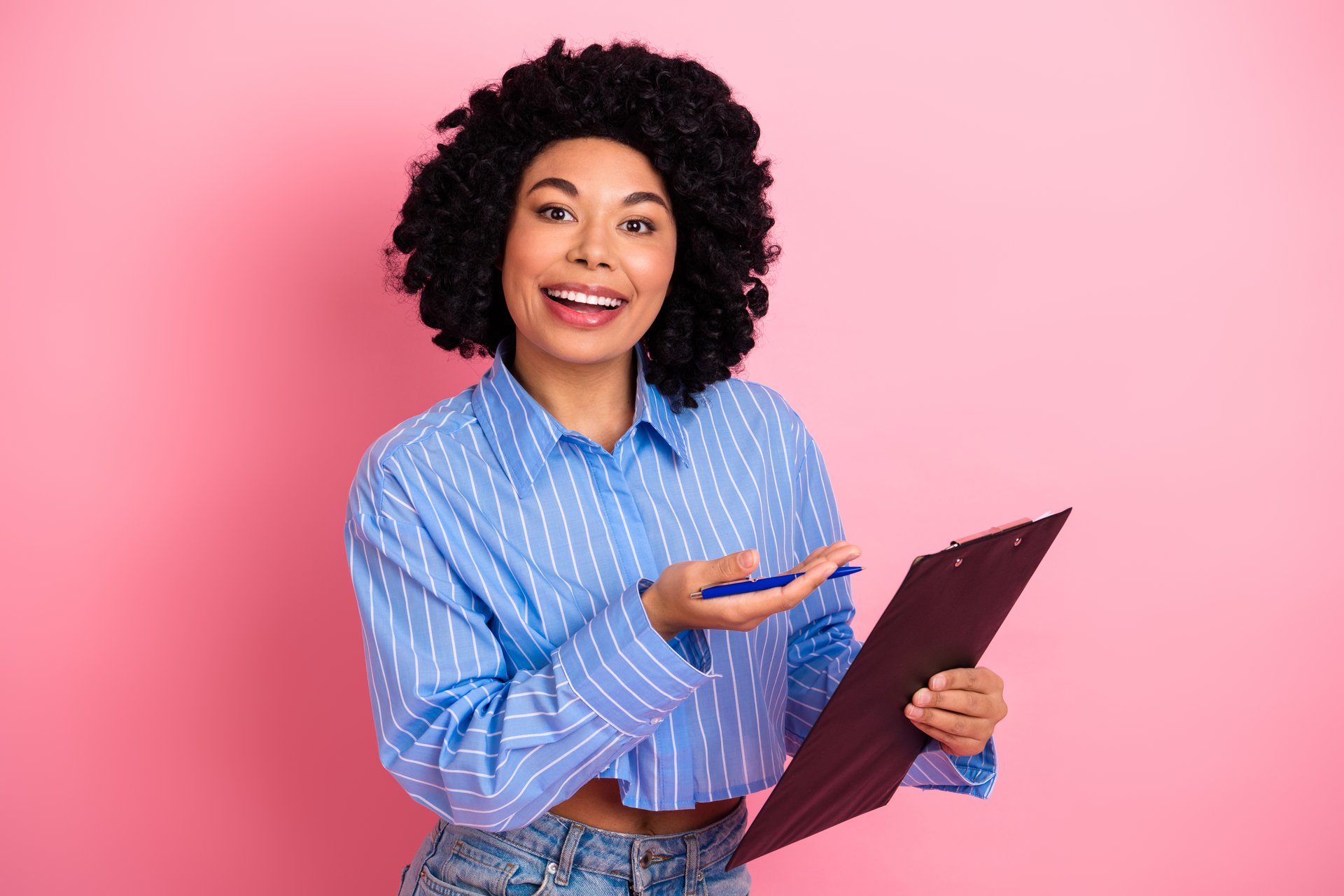 A cheerful young woman with curly hair, wearing a casual striped shirt, holding a clipboard, and gesturing confidently against a soft pink backdrop, conveying positivity and approachability.