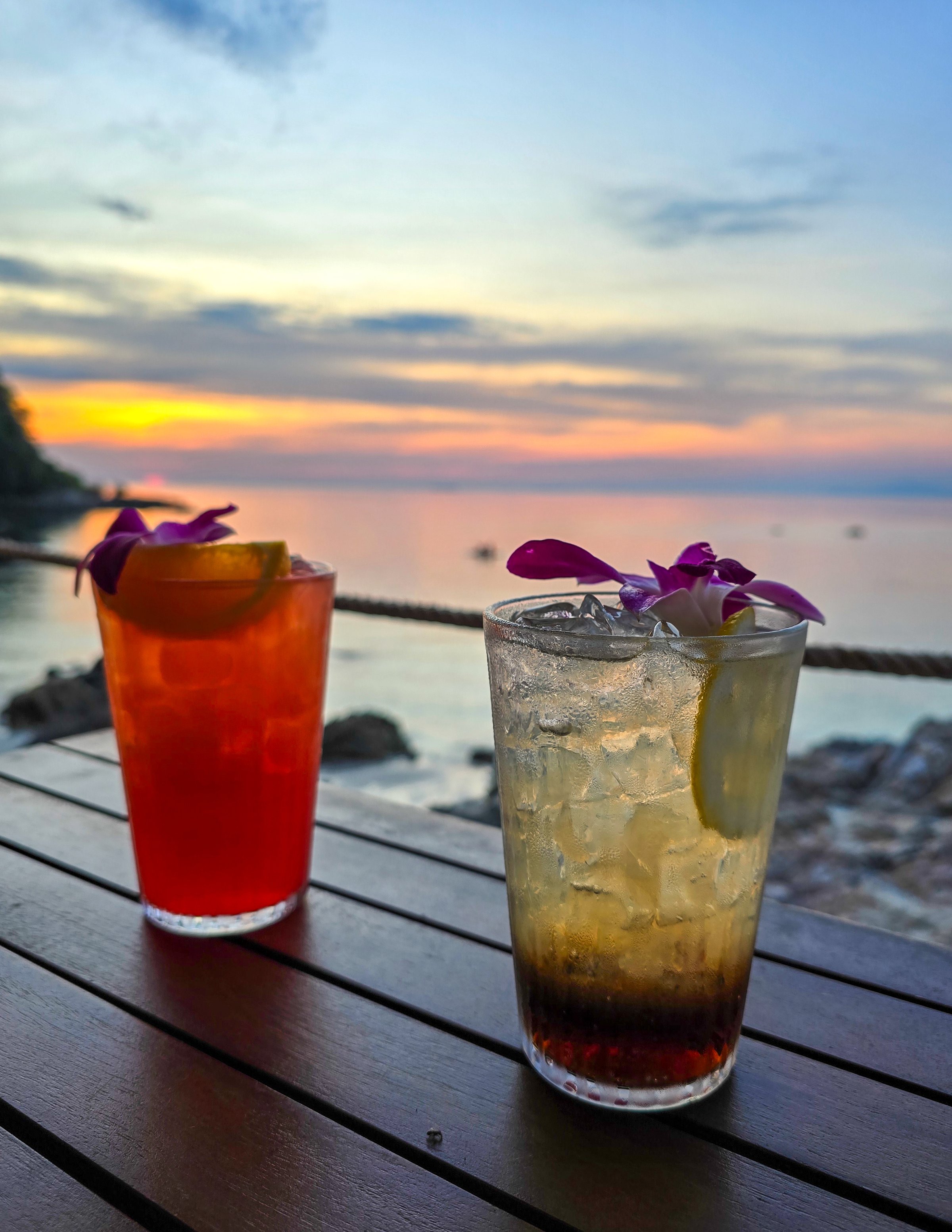 Two colorful cocktails sit on a wooden table, adorned with flowers, as the sun sets over the tranquil waters of Koh Munnork Island in Rayong Thailand.. A perfect evening escape awaits.