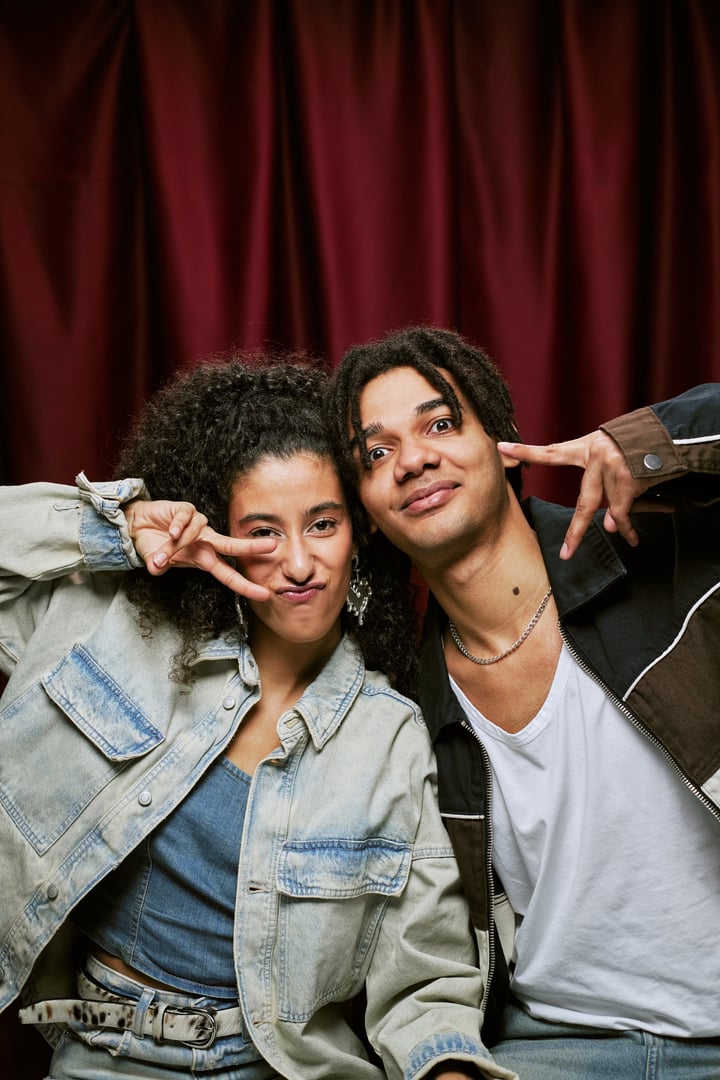 Young adult Black man and young adult multiethnic woman posing together in photo booth making peace signs with hands, smiling and looking directly at camera against red curtain backdrop