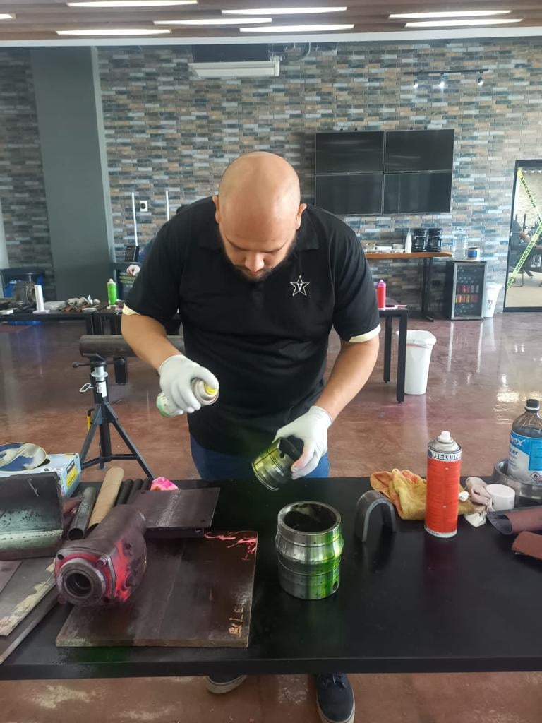 Man in gloves spray painting a metal object on a table with tools and cans in a workshop setting.