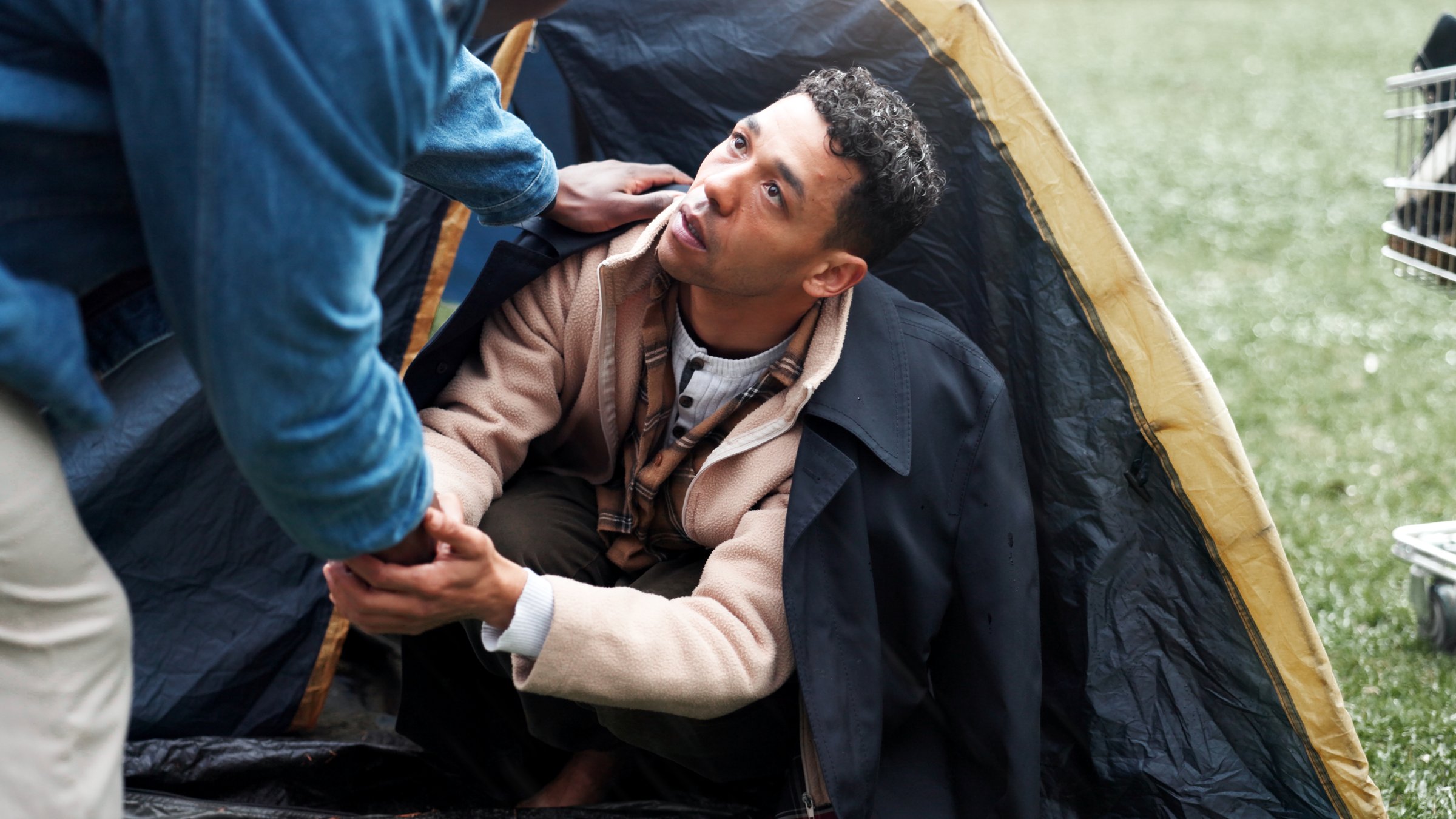 Man, poverty and homeless with tent, help and holding hands with support, kindness and care on ground. People, volunteer and hope with social awareness, connection and empathy on grass at park
