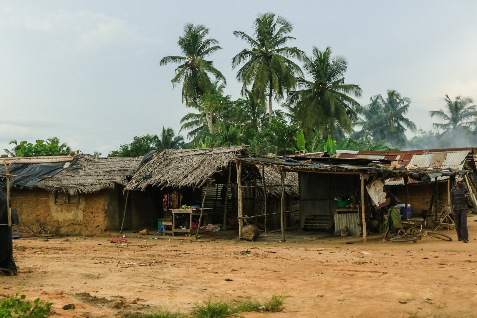 A rural village in Ivory Coast features mud houses, thatched roofs, and tall palm trees amidst a cloudy atmosphere.