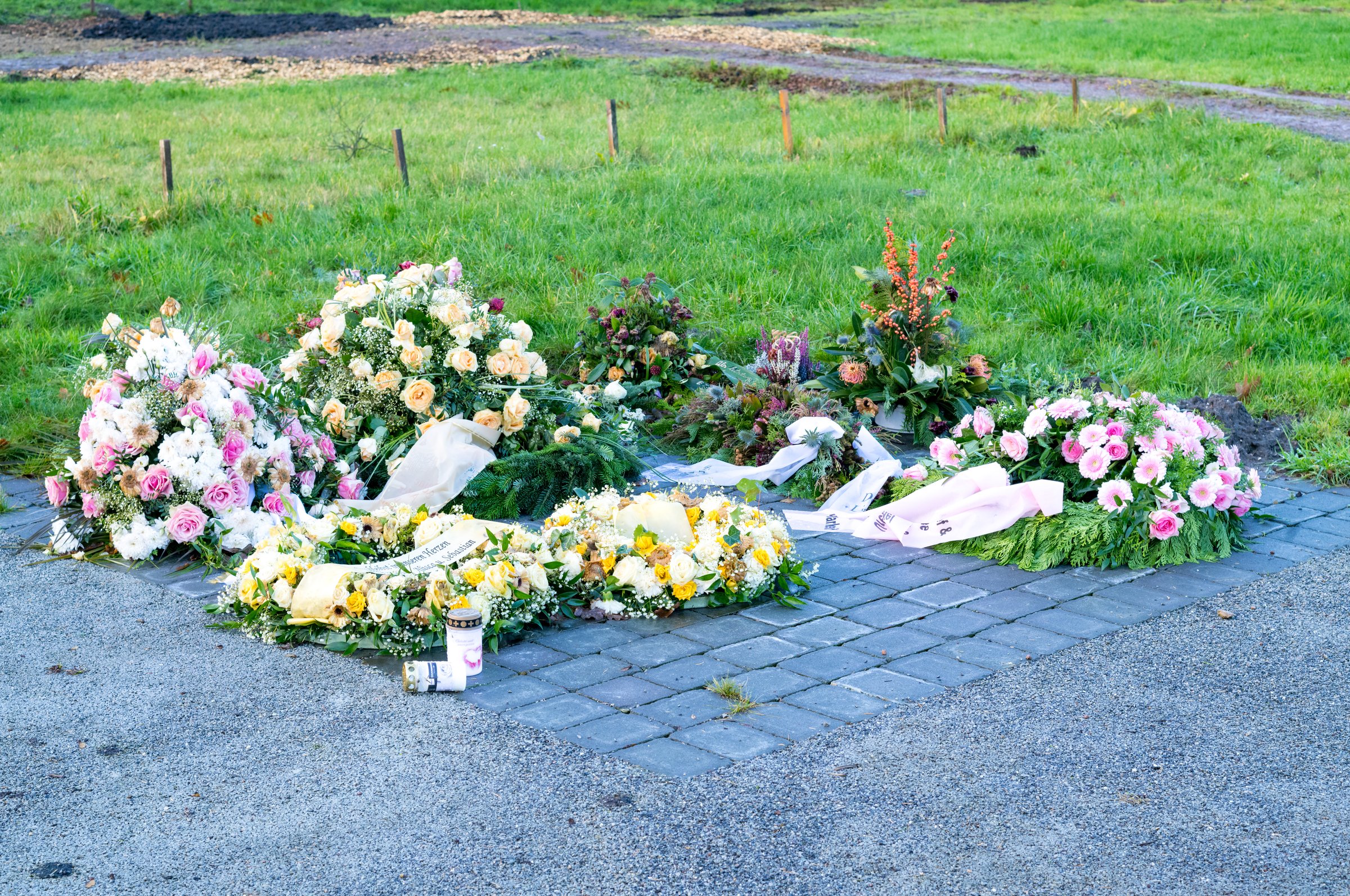 Colorful flower arrangements and wreaths commemorating a person lie on a paved area near a green lawn
