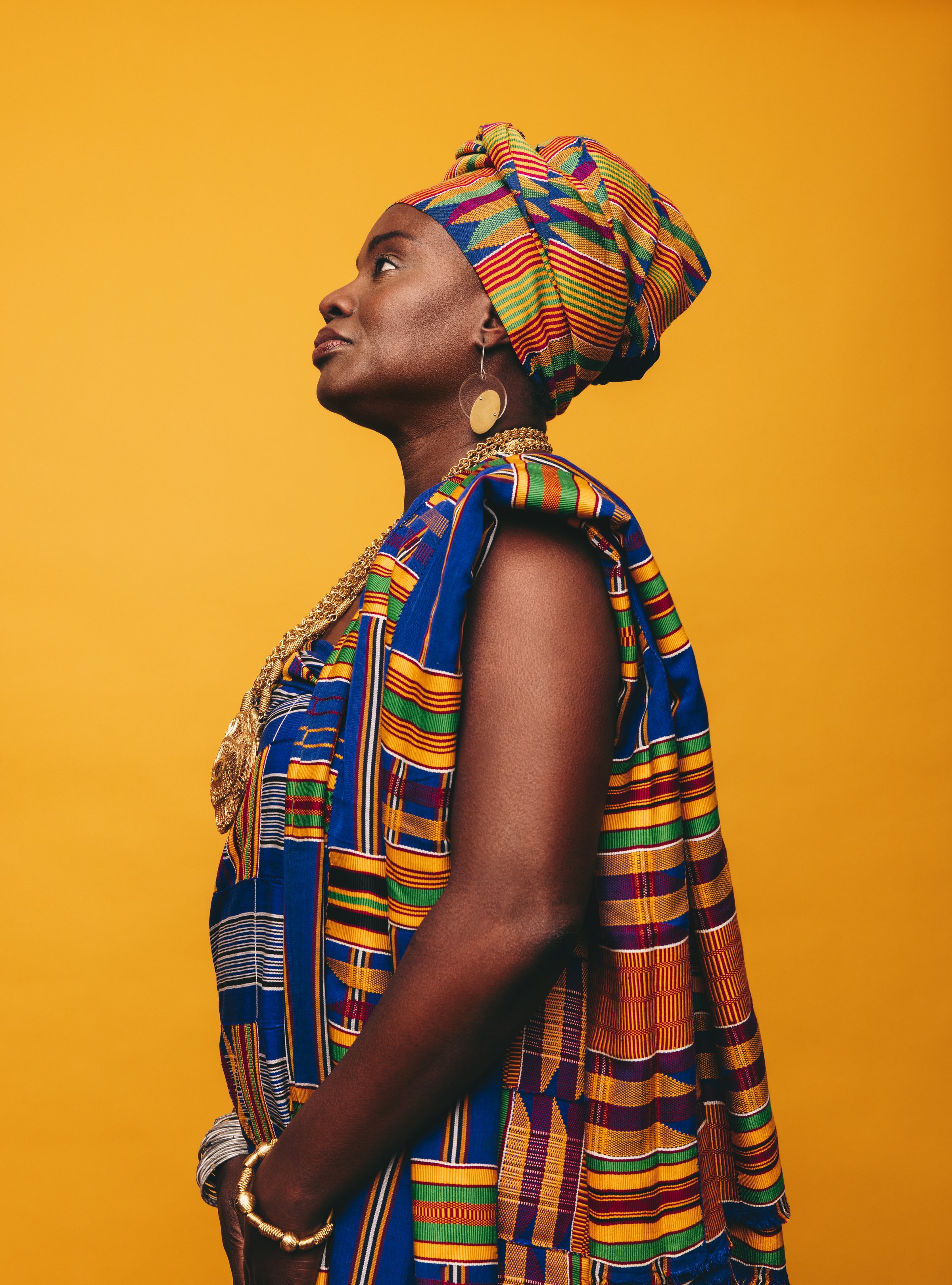 Elegant African woman wearing a traditional attire against a yellow background. Mature black woman dressed in colourful Kente cloth and golden jewellery. Confident woman embracing her rich culture.