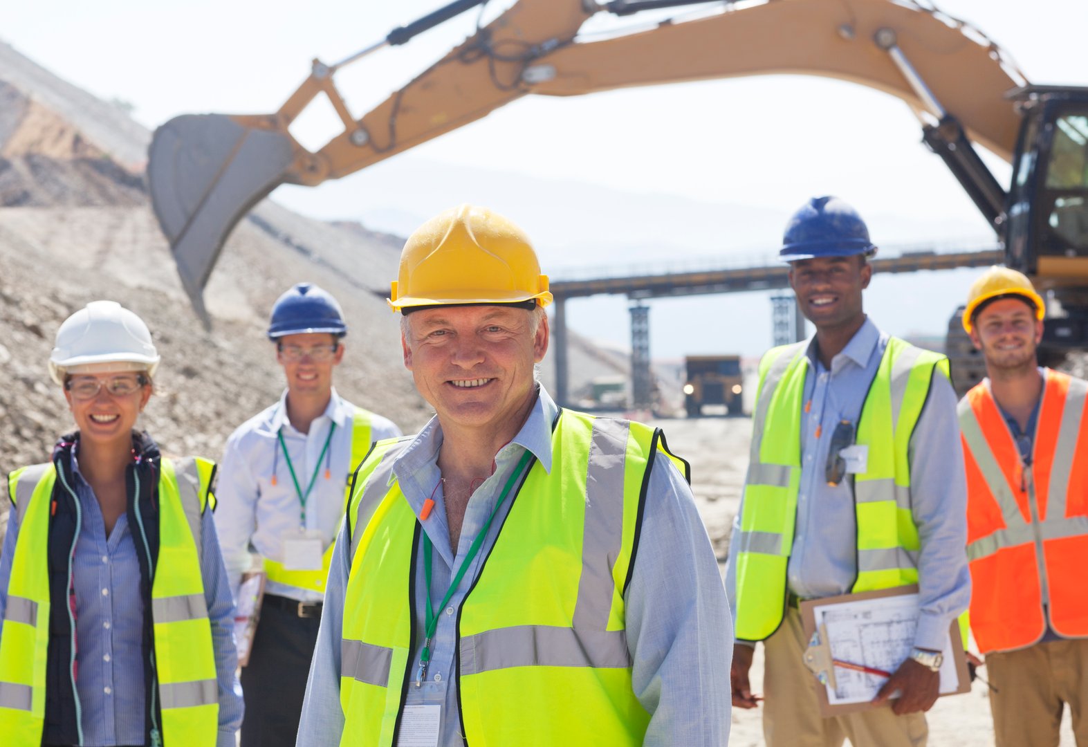 Construction workers wearing safety gear and hard hats stand in front of heavy machinery on a construction site.
