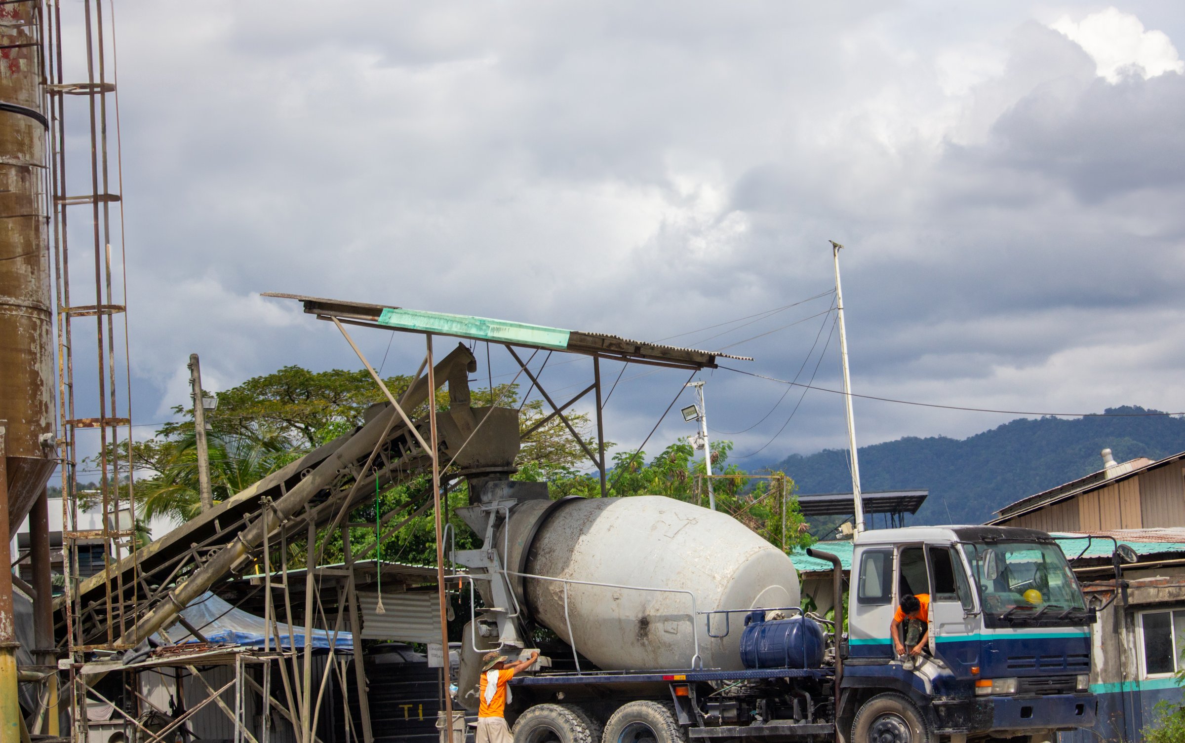 Concrete mixer truck positioned under a loading chute at a batching plant, surrounded by industrial structures and greenery. The large drum is mounted on a blue truck. Two workers wearing safety helmets and vests are present. Background features a cloudy sky and distant mountain range, creating an industrial landscape. The scene highlights construction processes and industrial activity.